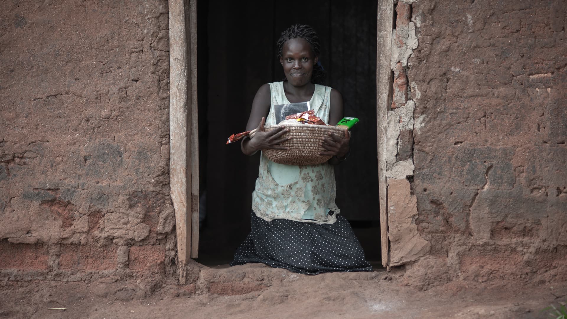 An elderly woman stands in a mud house doorway holding a basin.
