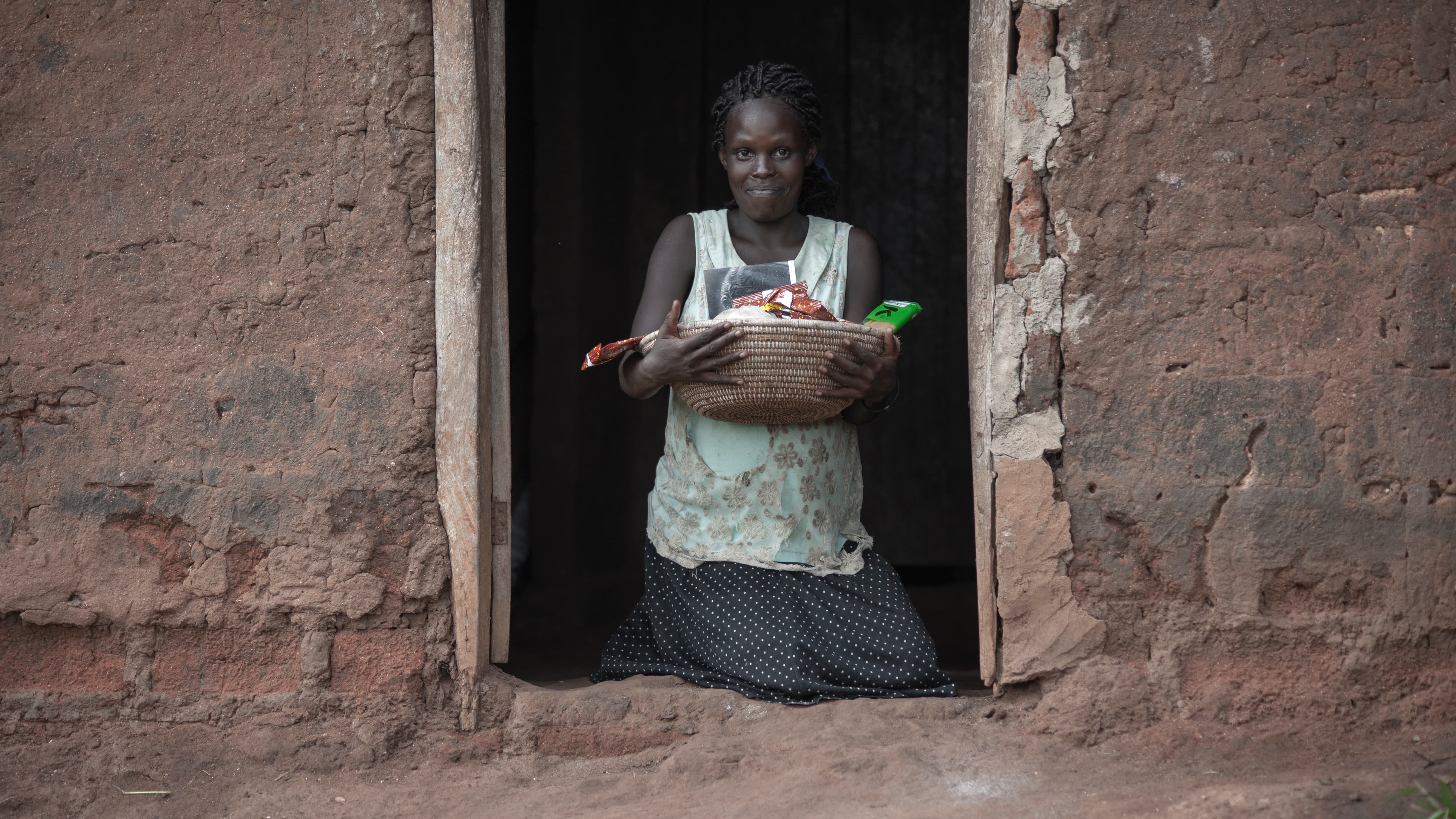 An elderly woman stands in a mud house doorway holding a basin.
