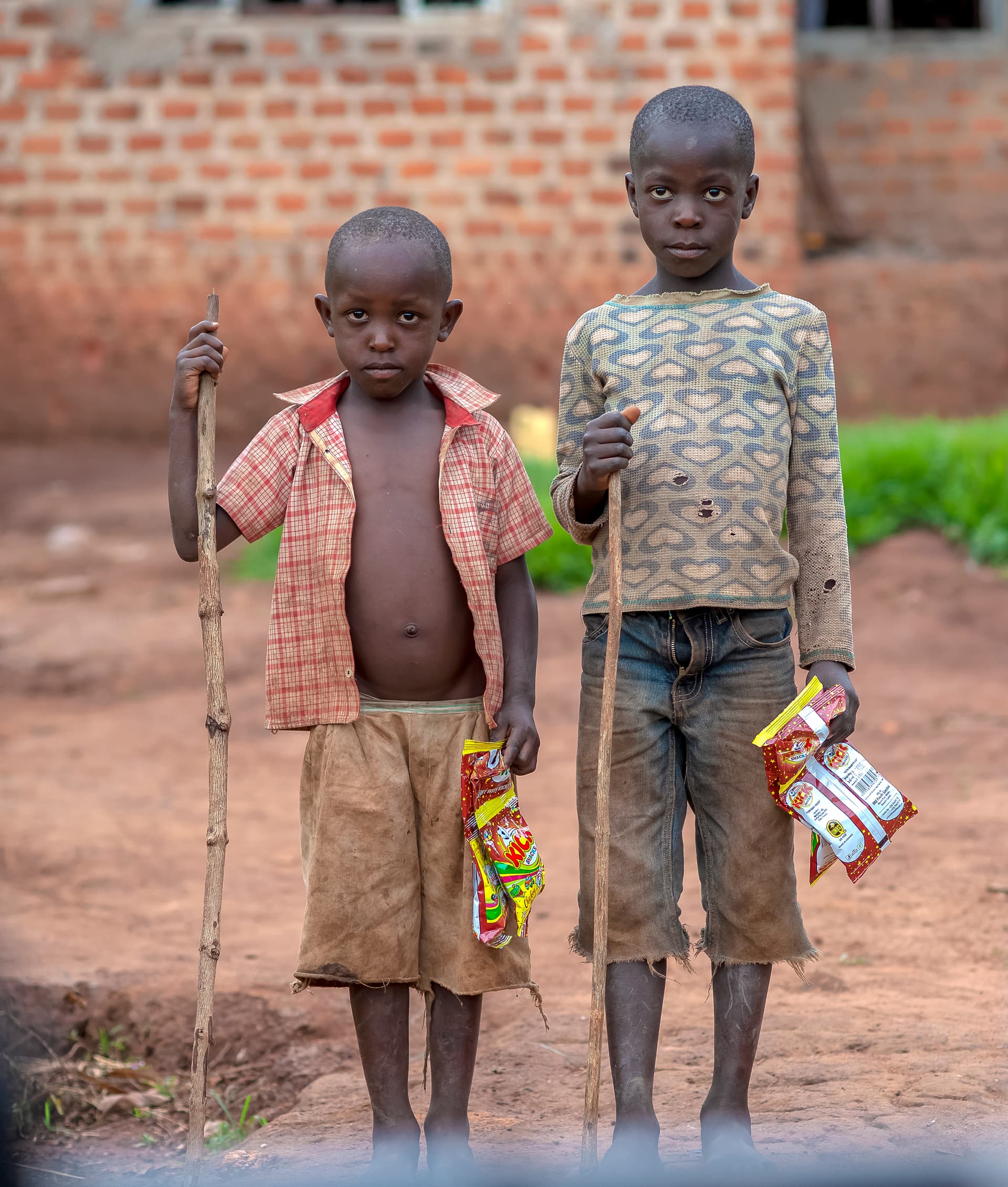 The same two boys stand together outdoors holding walking sticks and small Christmas food packages.