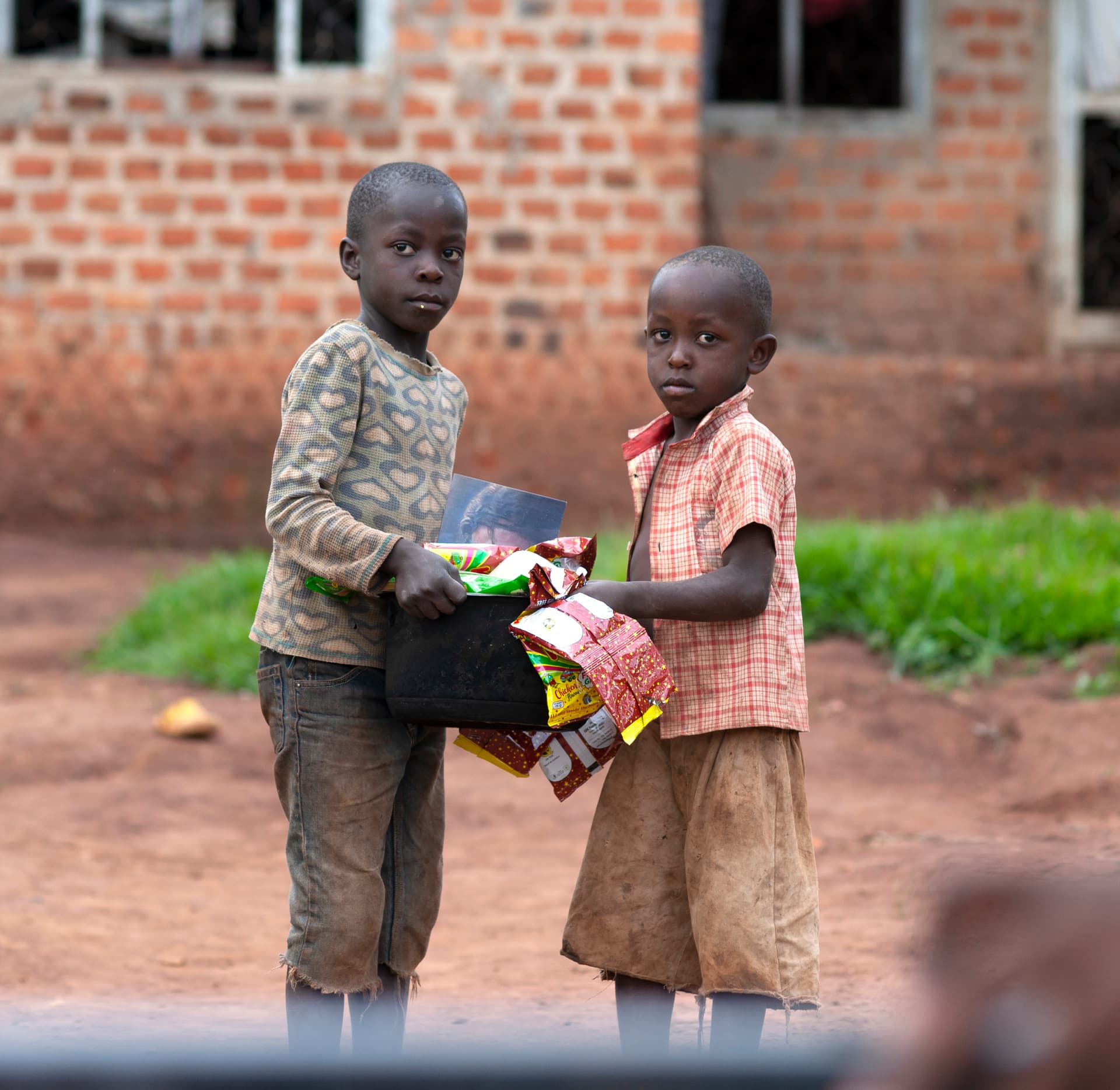 Two boys stand together outside a brick building holding a pot of food gifts.