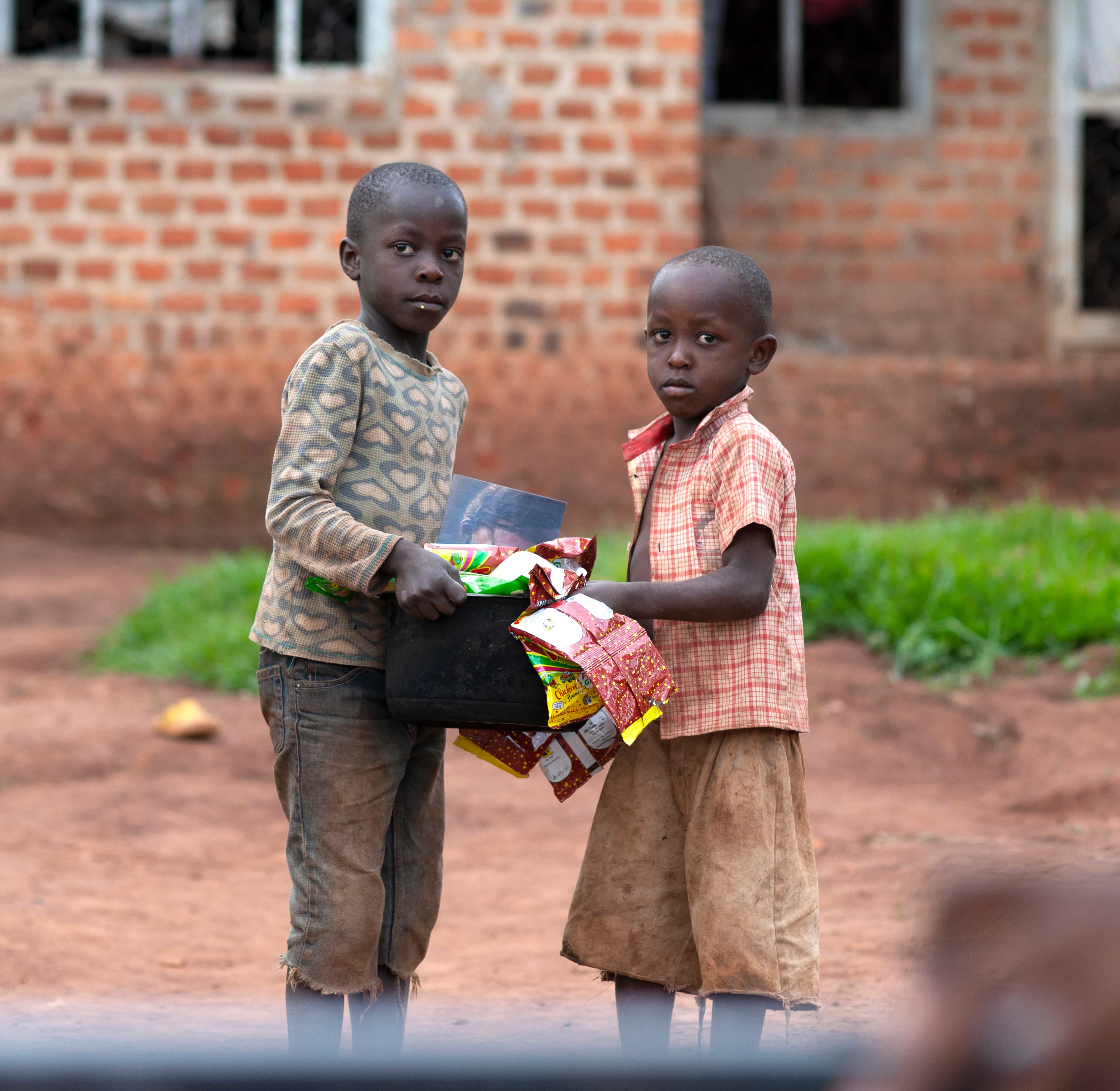 Two boys stand together outside a brick building holding a pot of food gifts.
