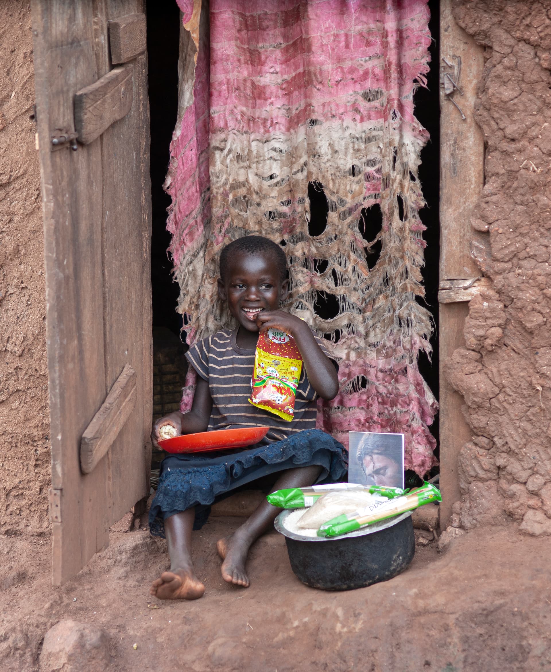 A child sits in a curtained doorway holding a food gift bag beside a metal basin.