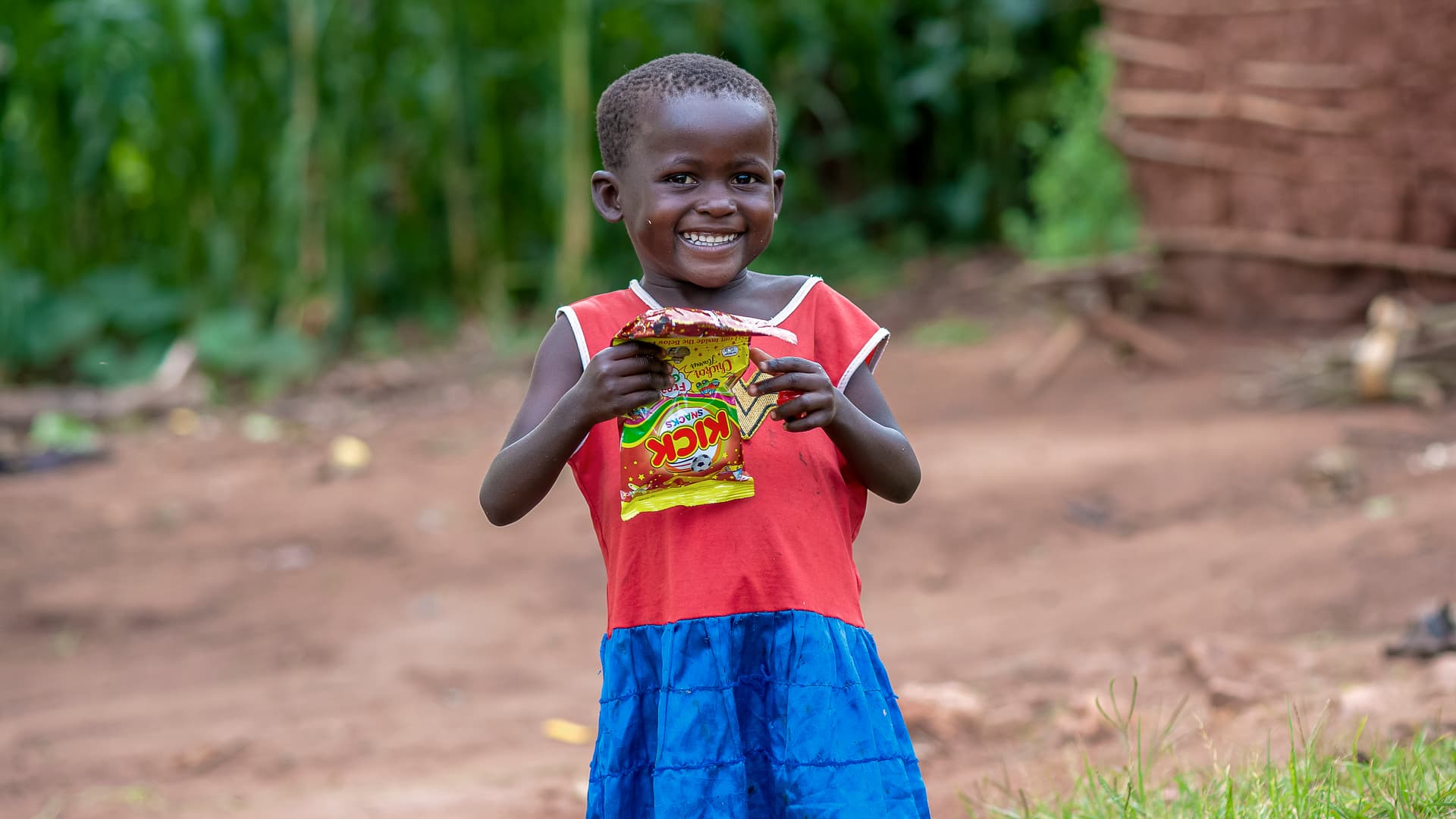 A child in a red and blue outfit smiles while holding a food gift bag.