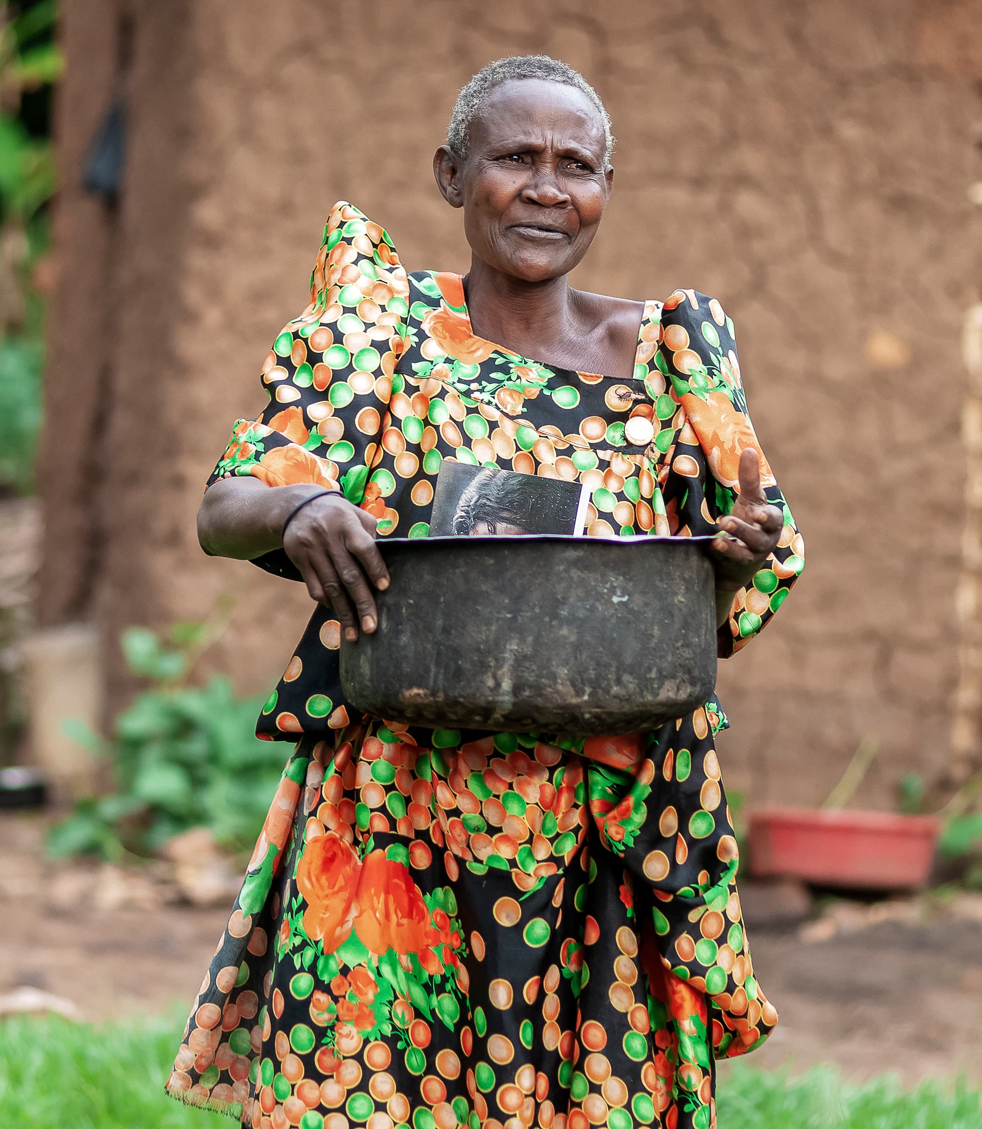 An elderly woman in a colorful dress holds a large pot filled with food gifts.