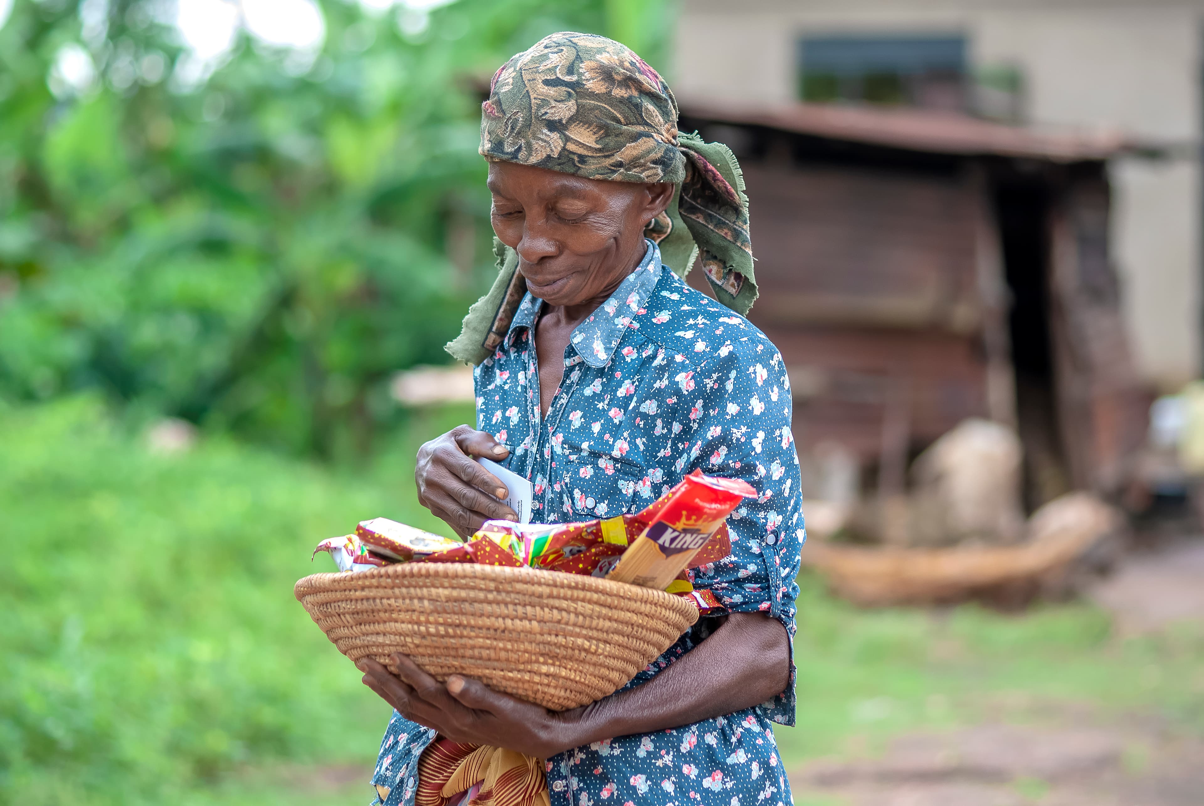 A woman in a patterned dress holds a basket of food gifts near a mud home.