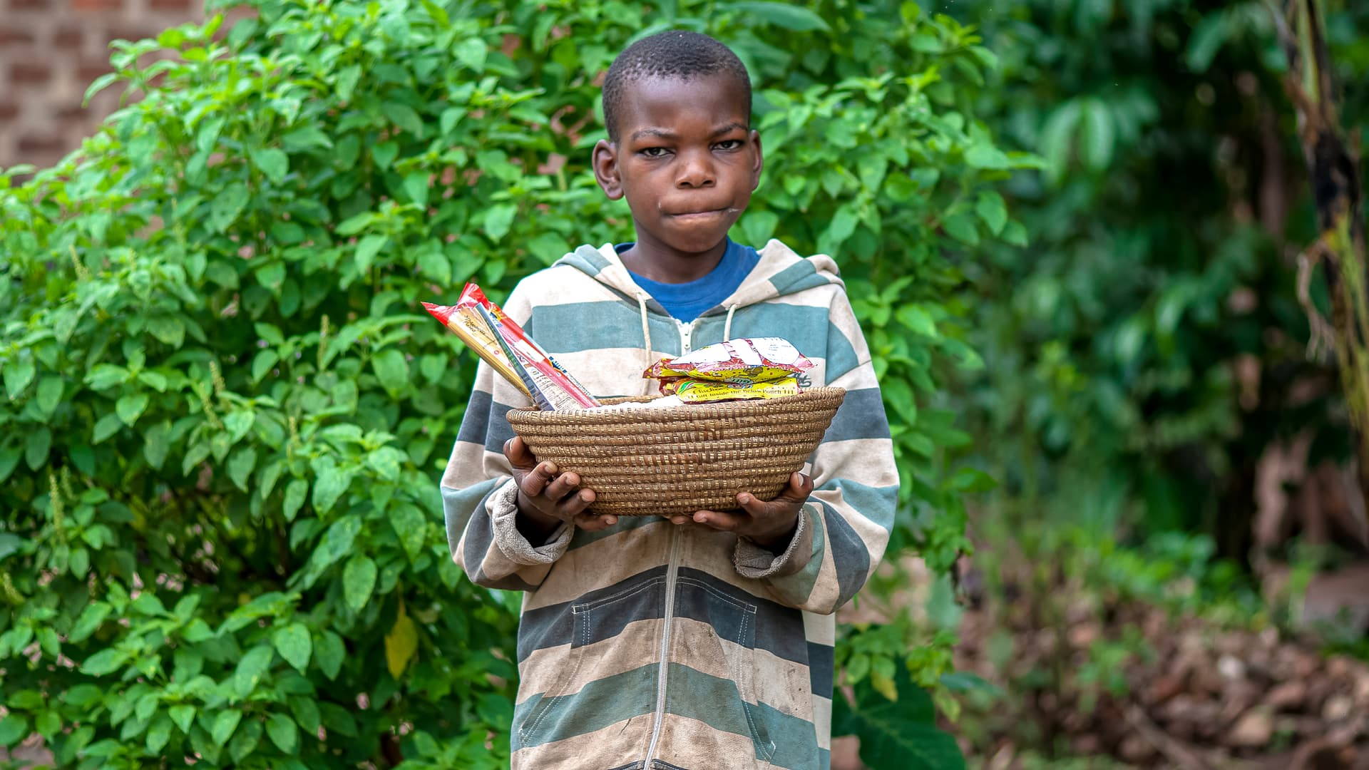 A boy holds a basket of food gifts while standing among green plants.