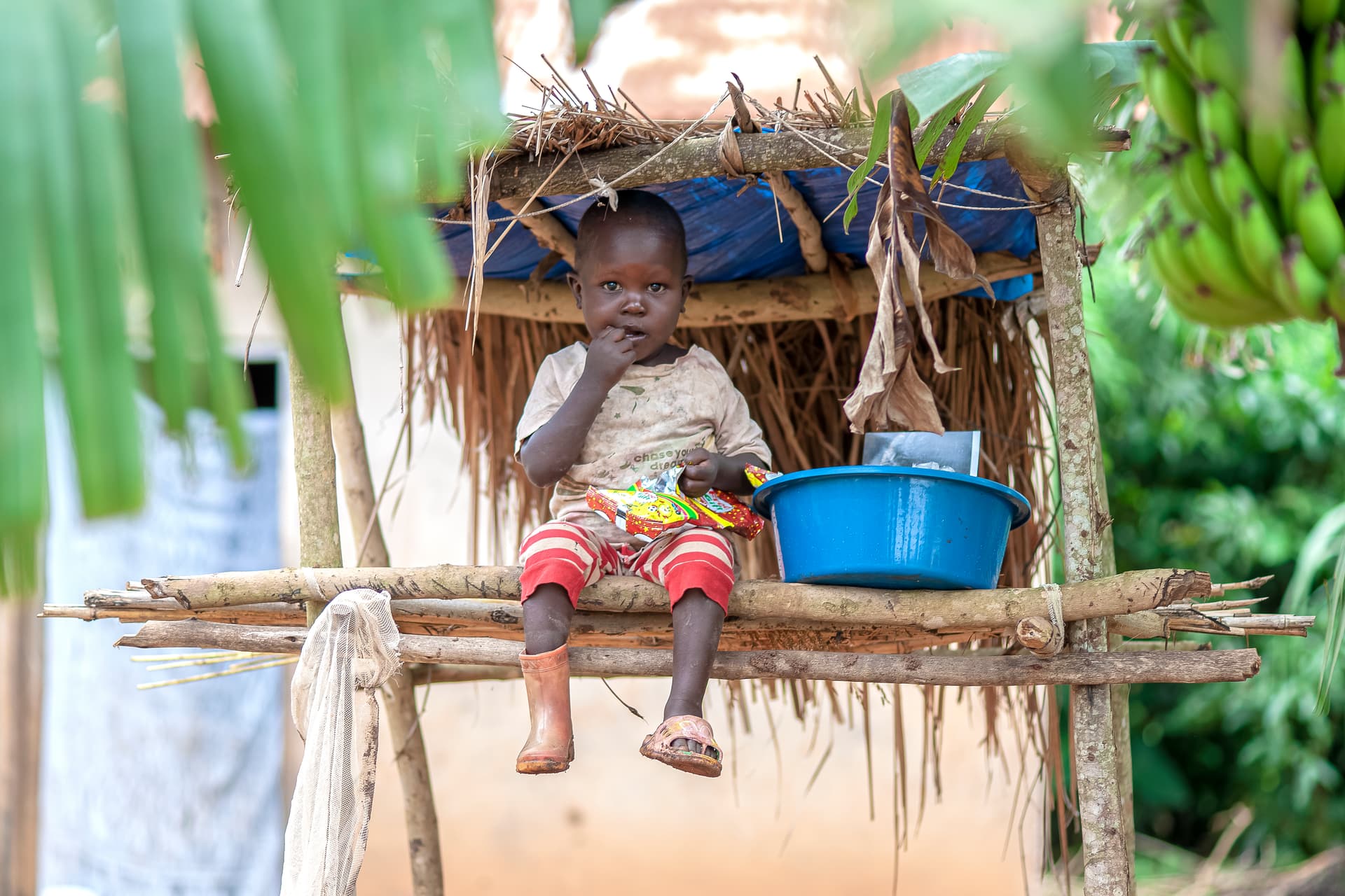 A child sits under a thatched-roof outdoor shelter beside a blue plastic basin.
