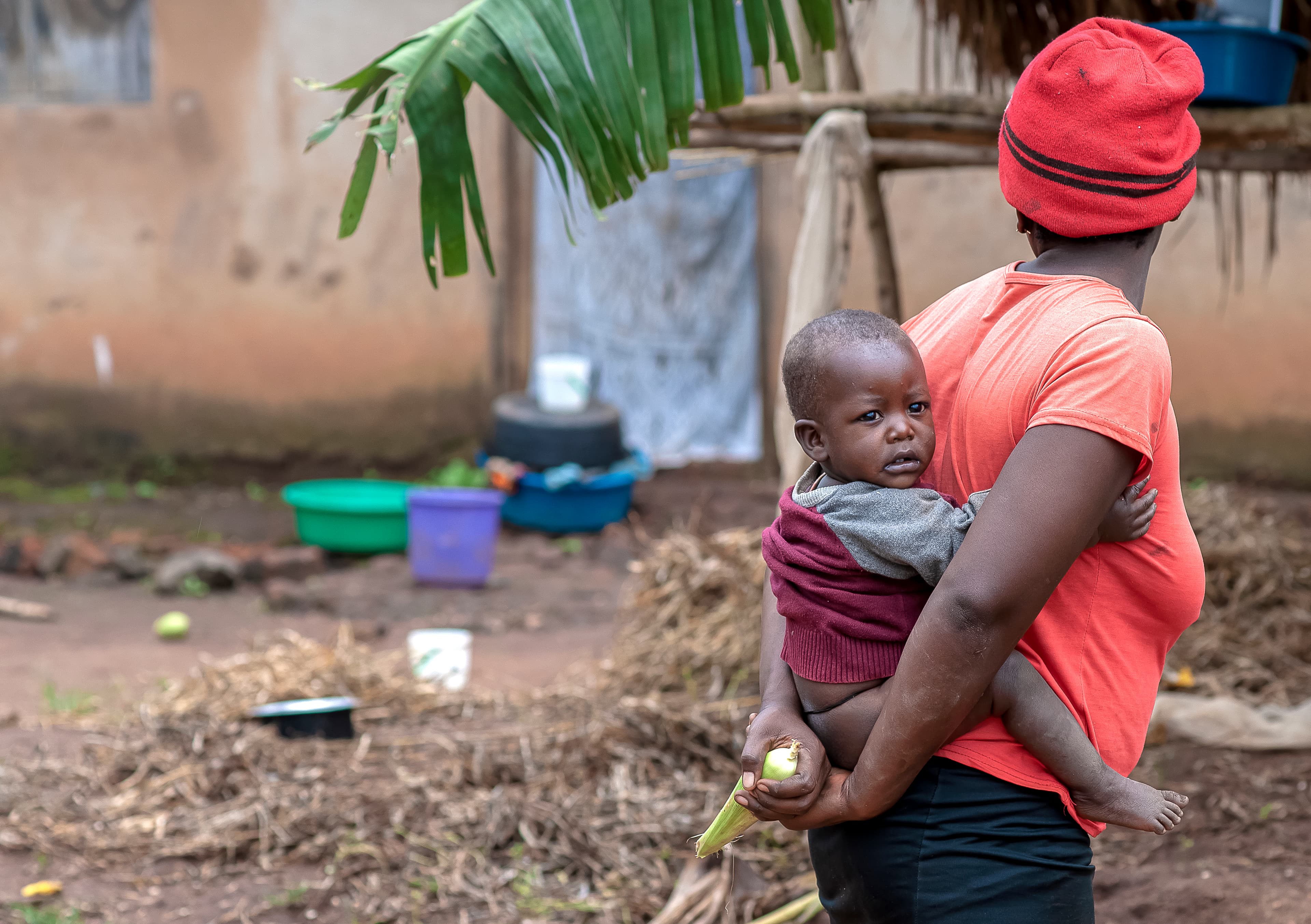 A woman in a red headscarf carries a baby on her back near household items.