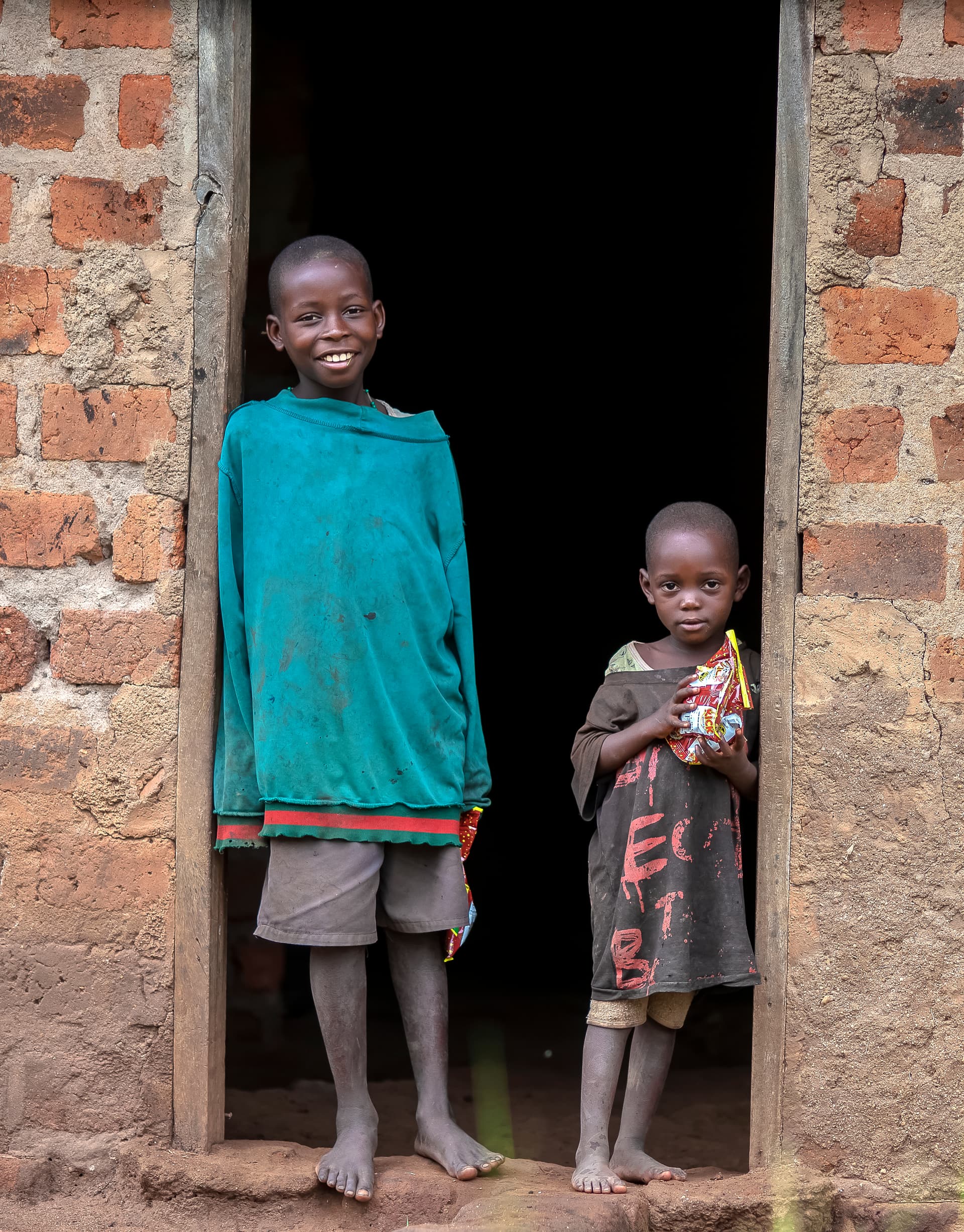 A taller boy and a younger child stand together in a doorway of a home.