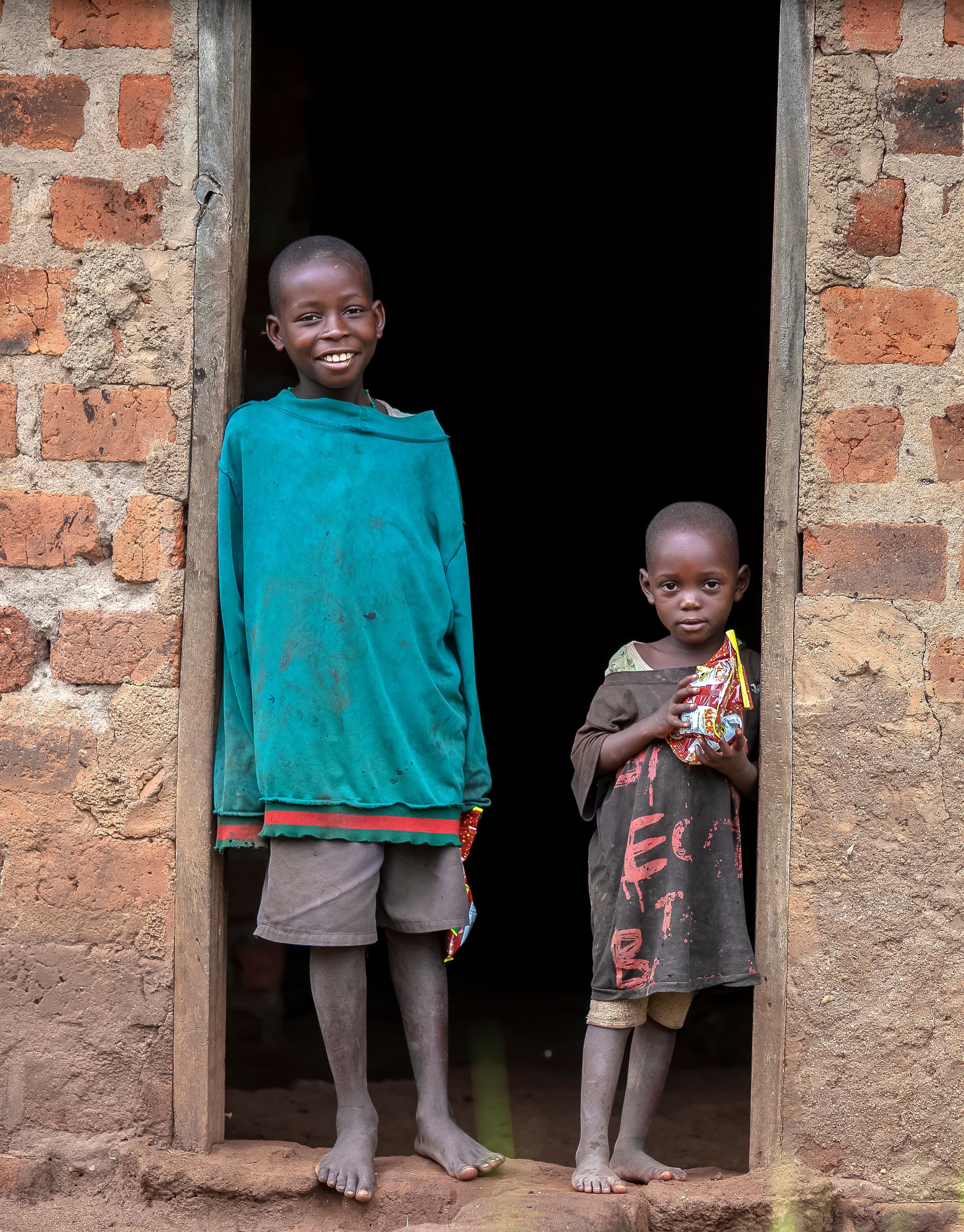 A taller boy and a younger child stand together in a doorway of a home.