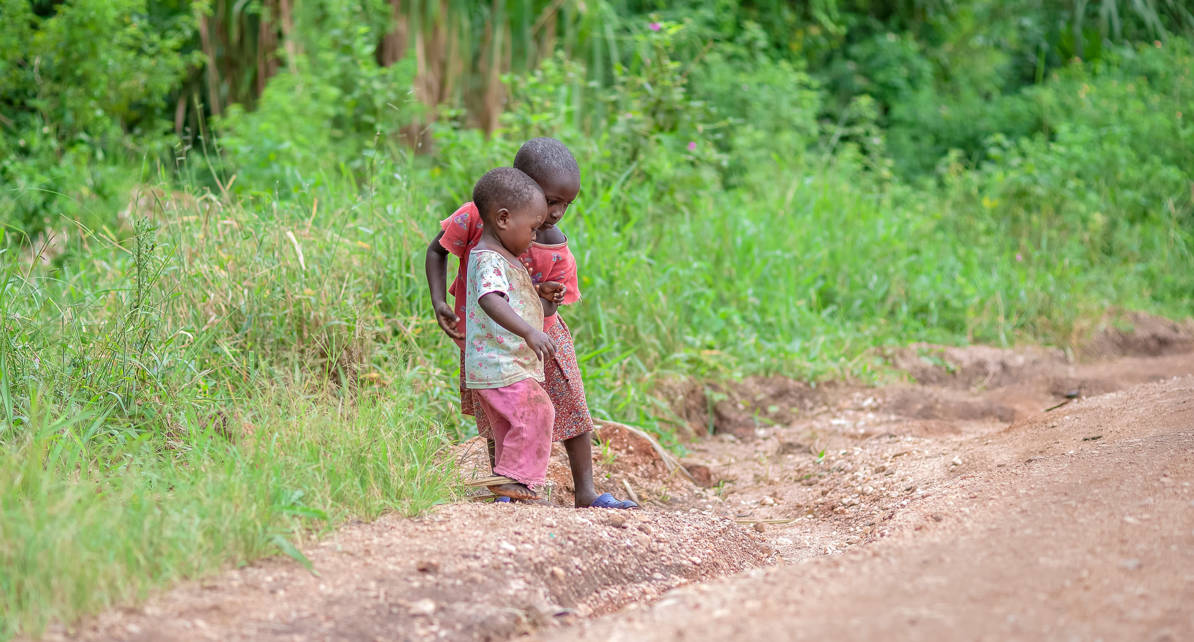 A child walks alone down a dirt road bordered by green fields, seen from a distance.