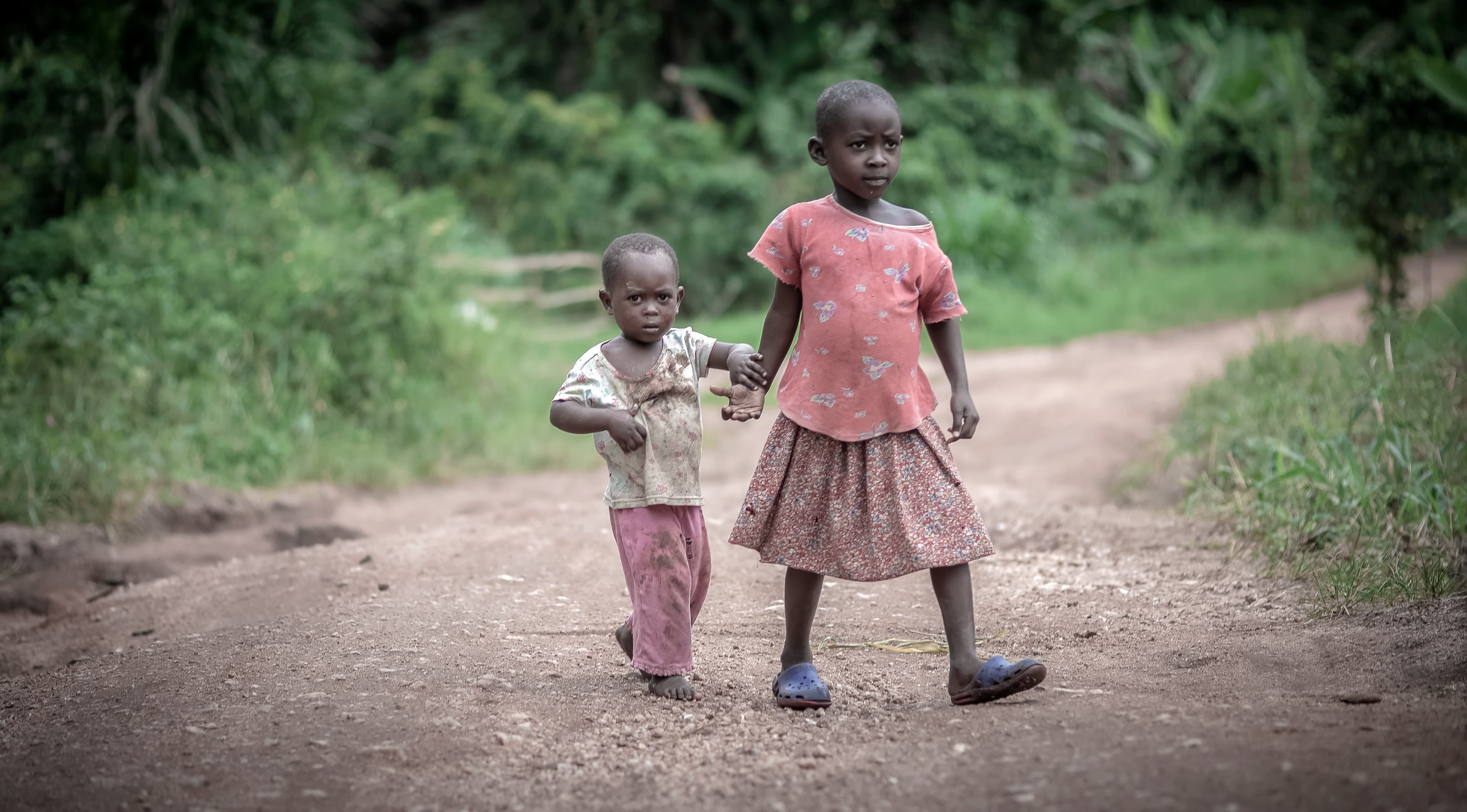 Two young children walk together along a dirt path near a village home.