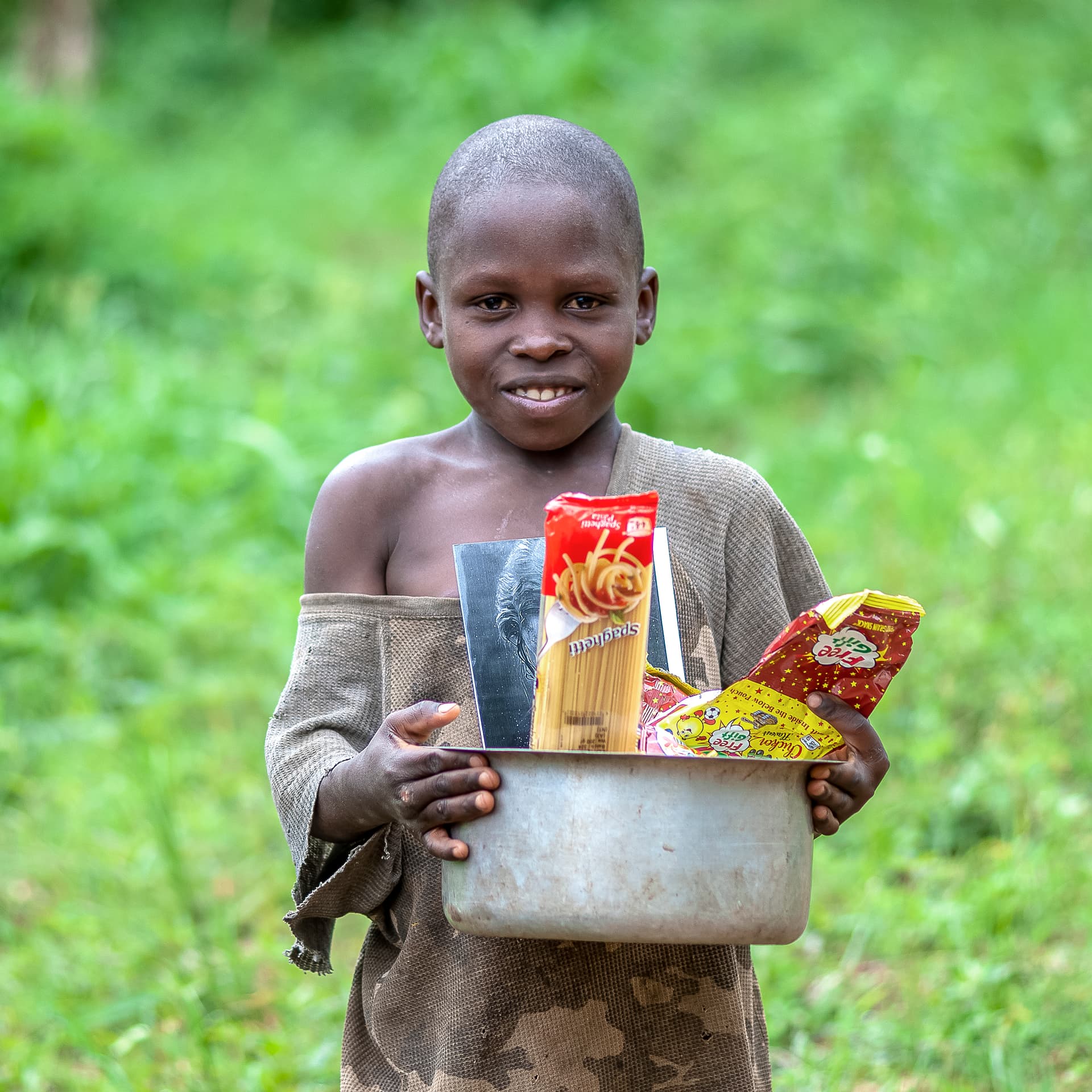 A child holds a metal basin filled with grocery items outdoors.