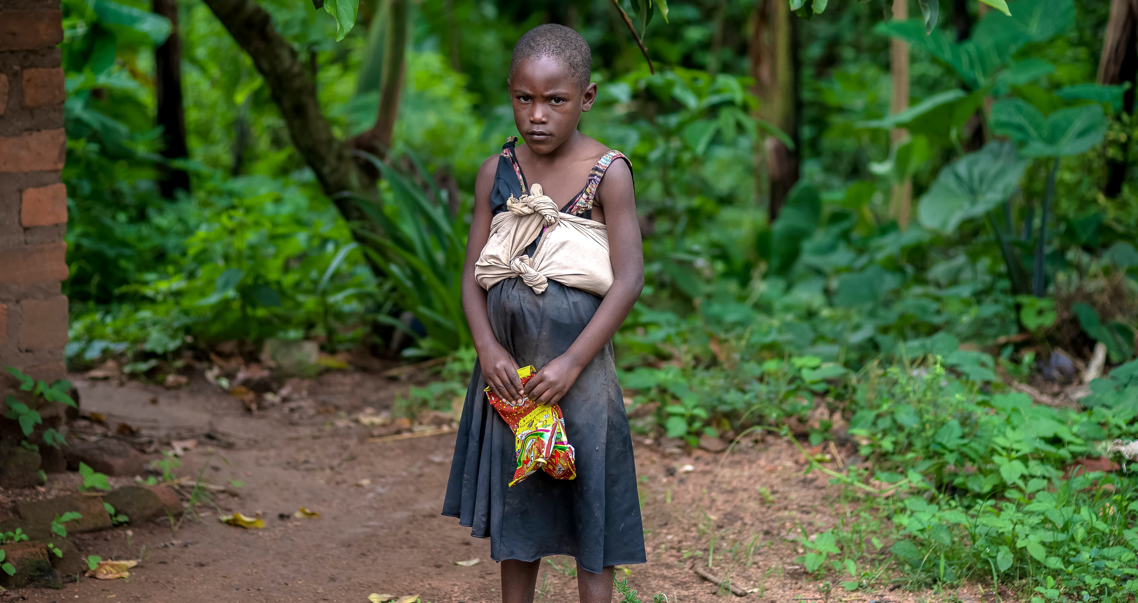 A young girl in a dark dress walks alone through a forested dirt path.