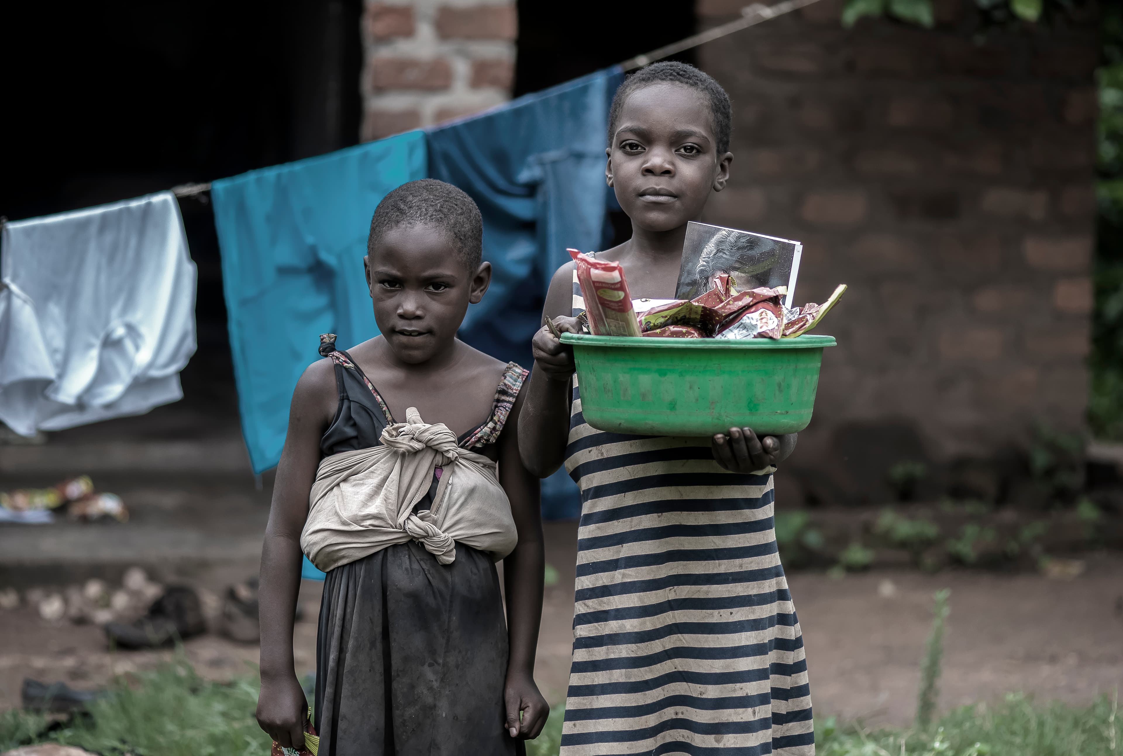Two children stand near a clothesline, one holding a green plastic basin of food.
