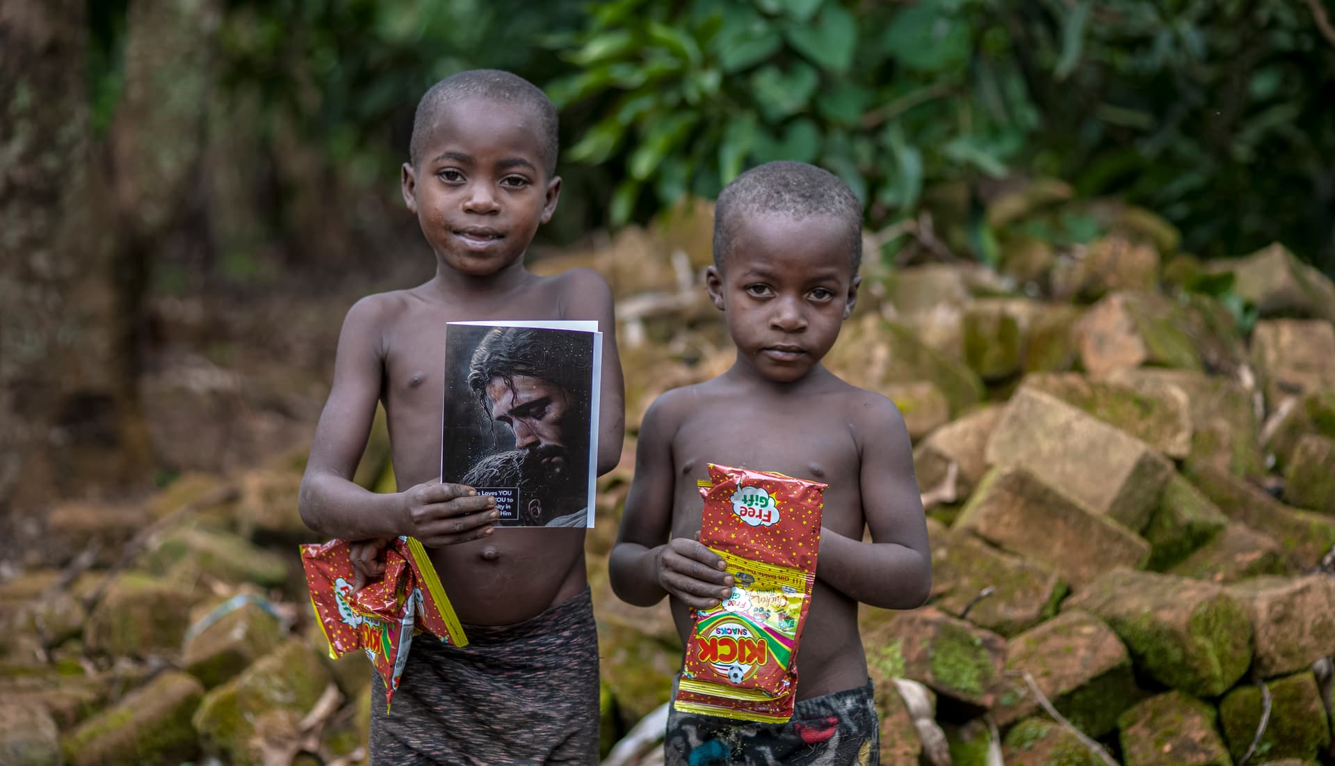 Two children hold a printed photo and food items near a pile of stacked bricks.