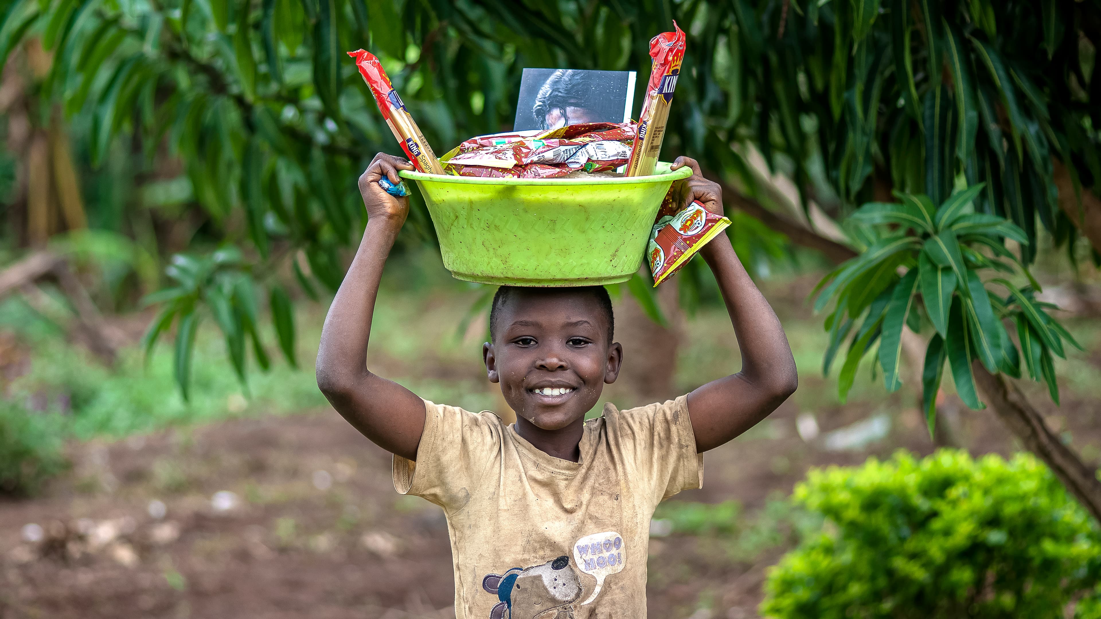 A boy lifts a green and yellow basin of food gifts above his head, smiling.