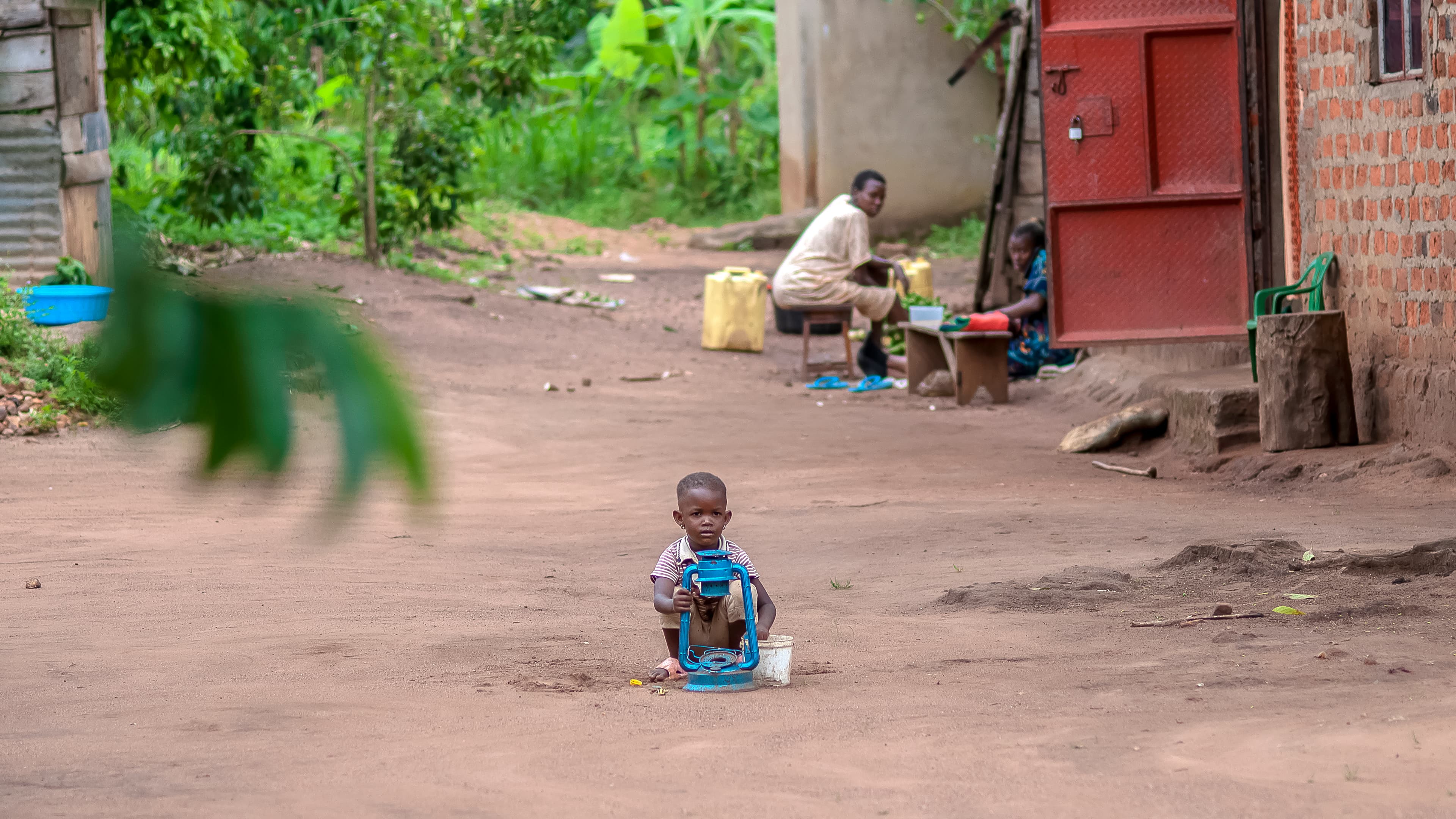 A child crouches alone on a dirt path near village buildings, seen from a distance.