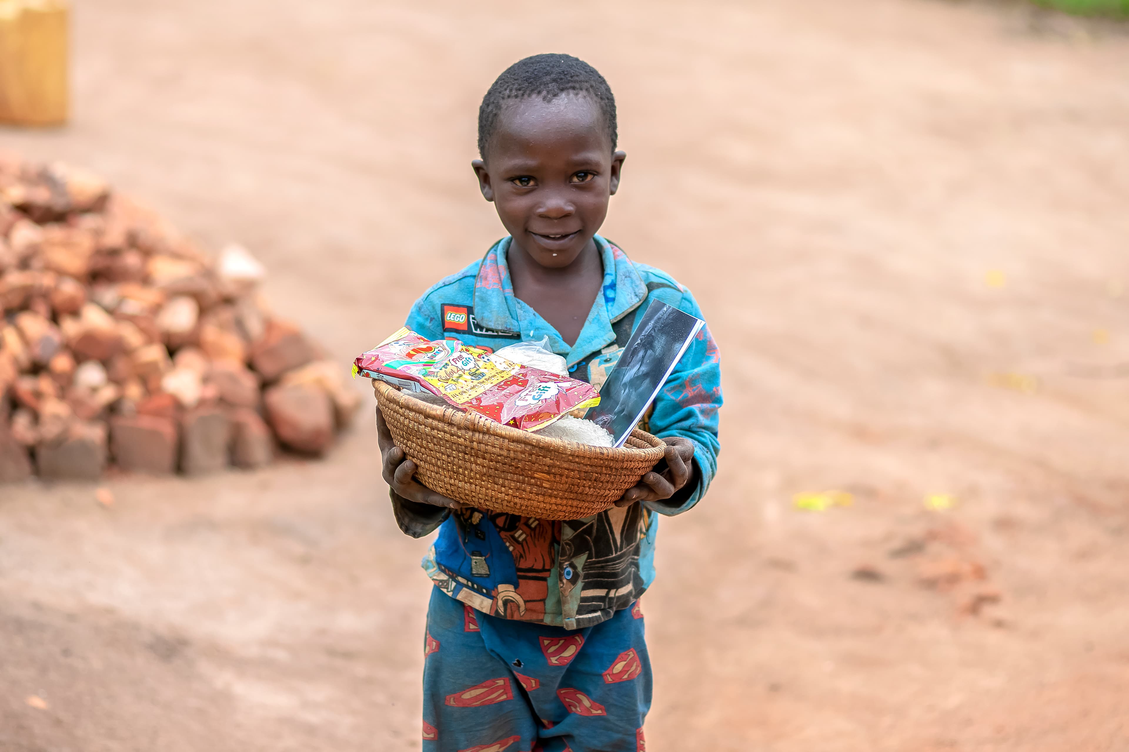 A boy carries a basket of food gifts while walking near piled bricks.