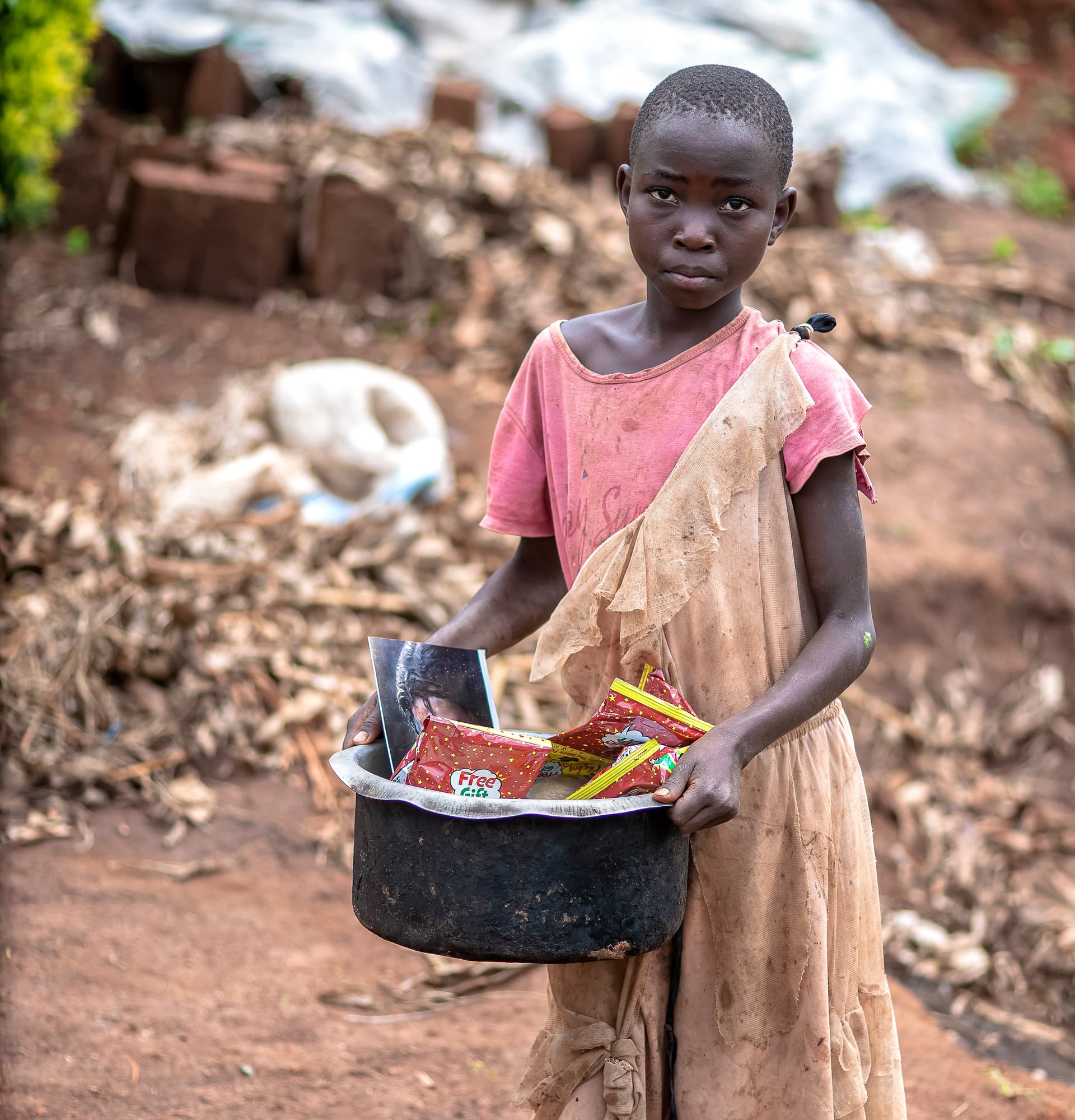 A girl holds a pot containing food gift items while standing near brick rubble.