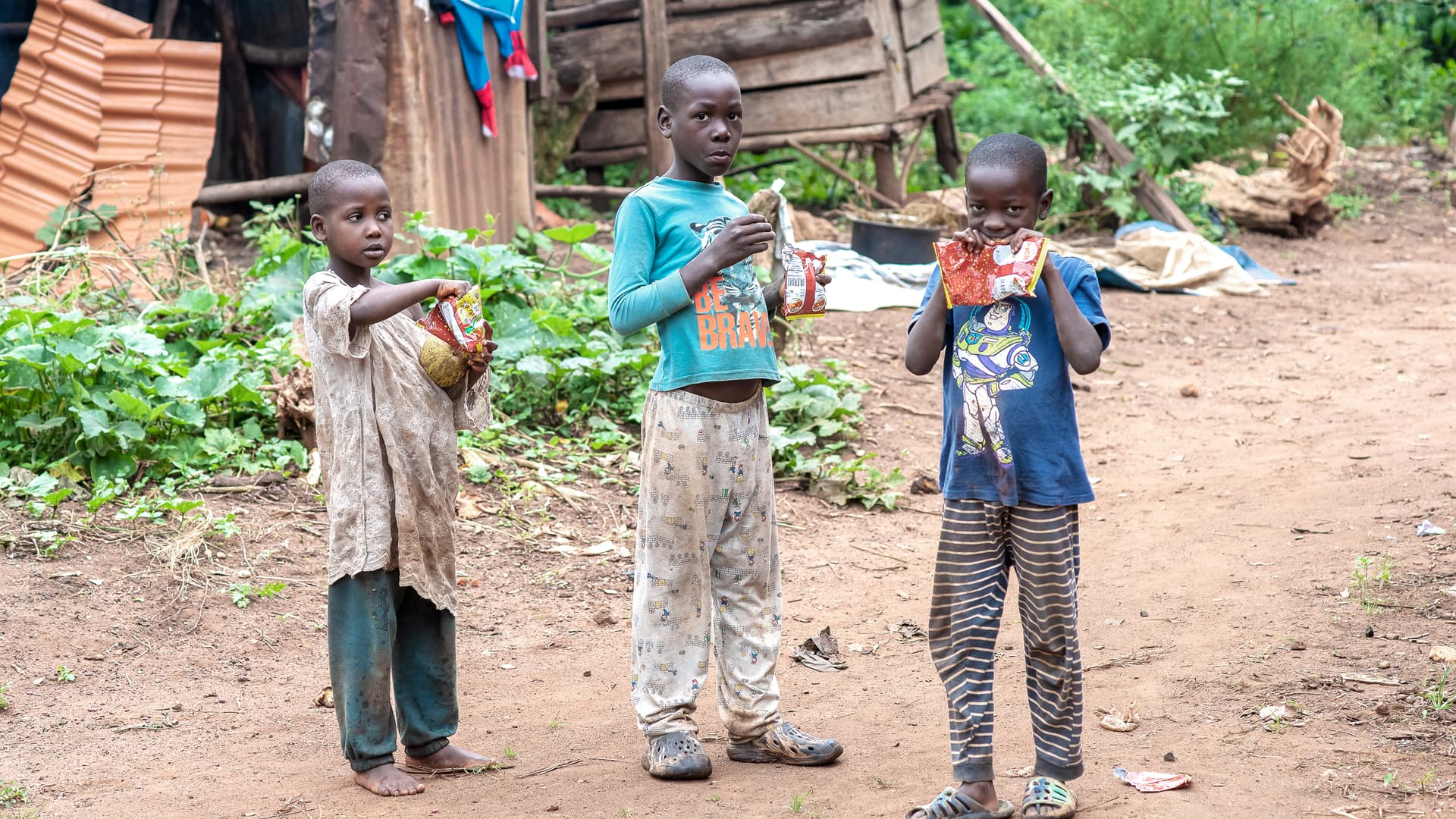A group of children stand outdoors near a rustic thatched shelter.