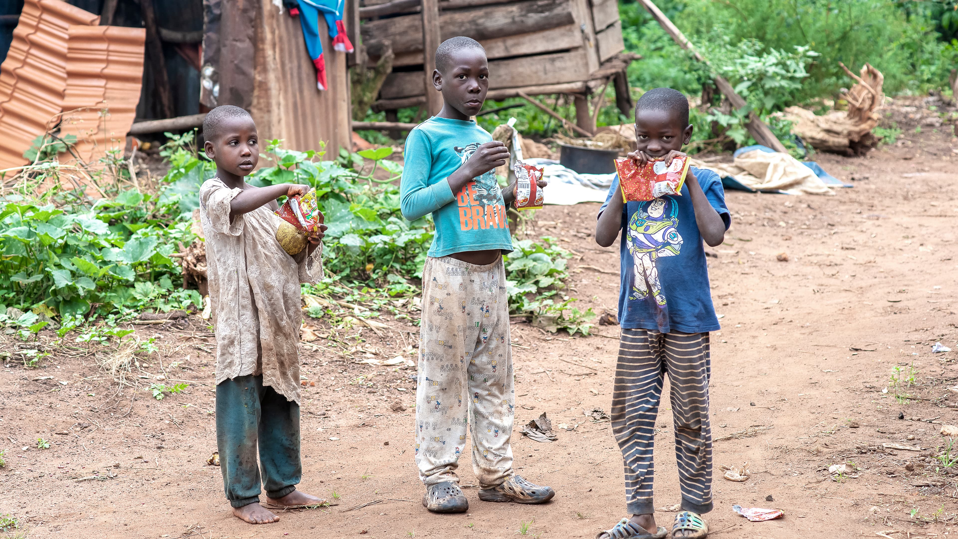 A group of children stand outdoors near a rustic thatched shelter.