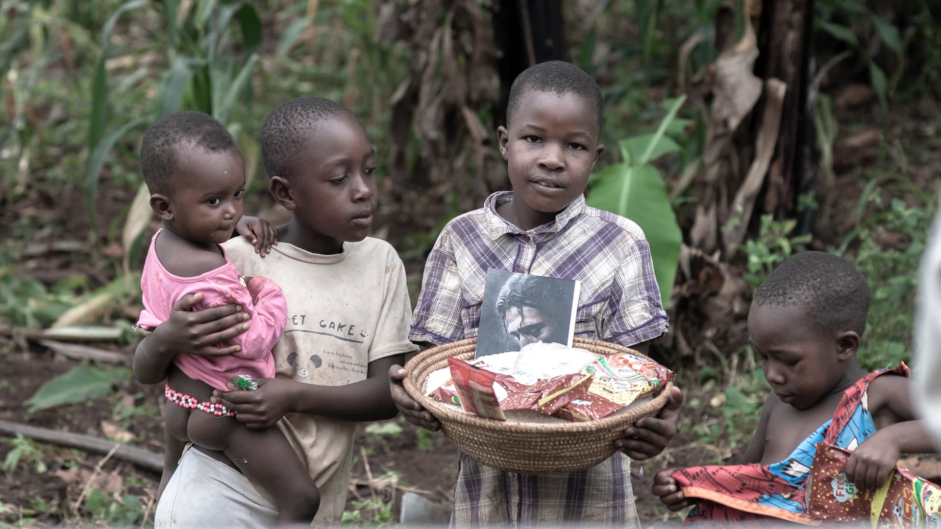A group of children stand together outdoors, one carrying a toddler.