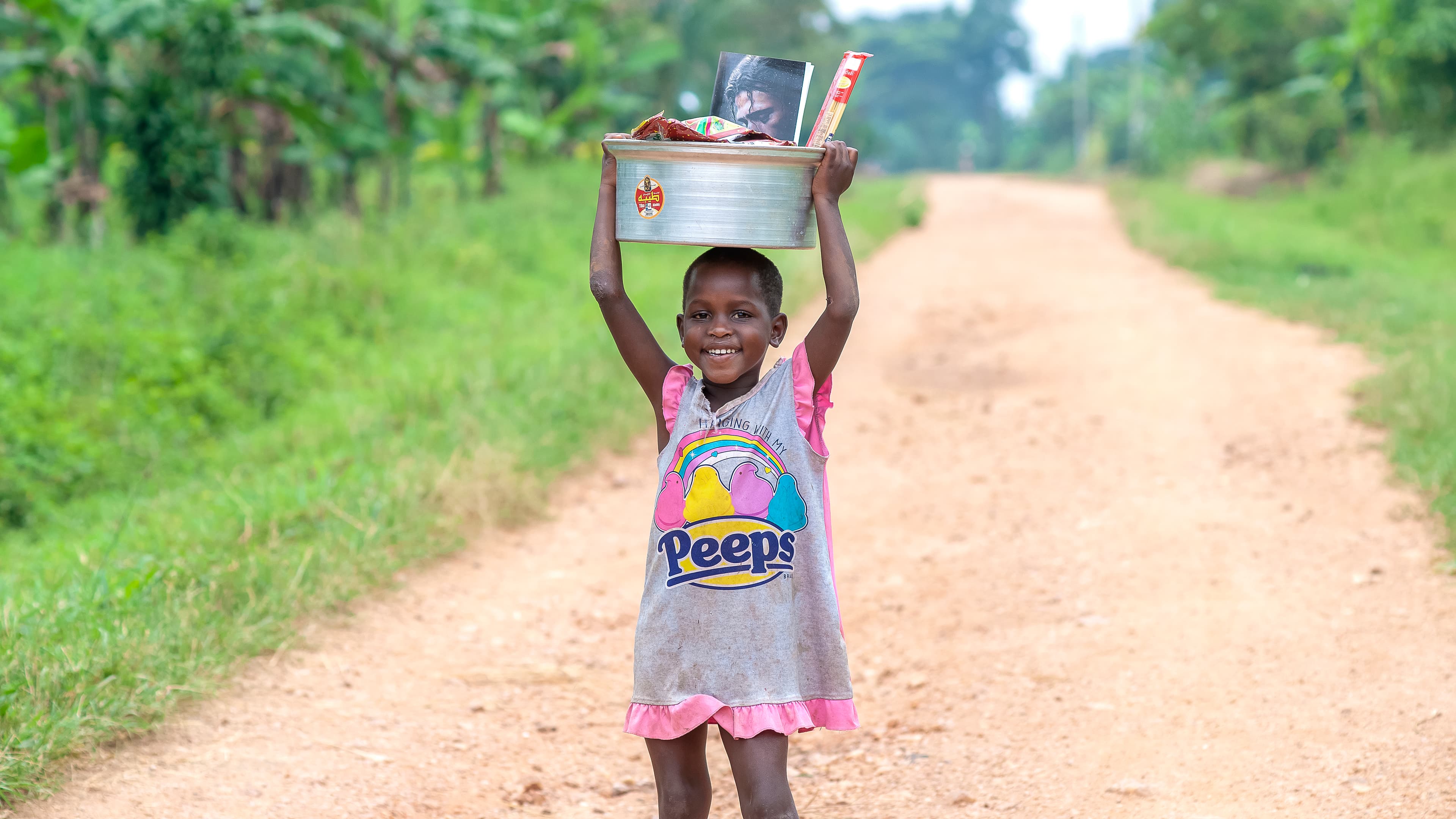 A girl carries a metal pot balanced on her head while walking along a dirt road.