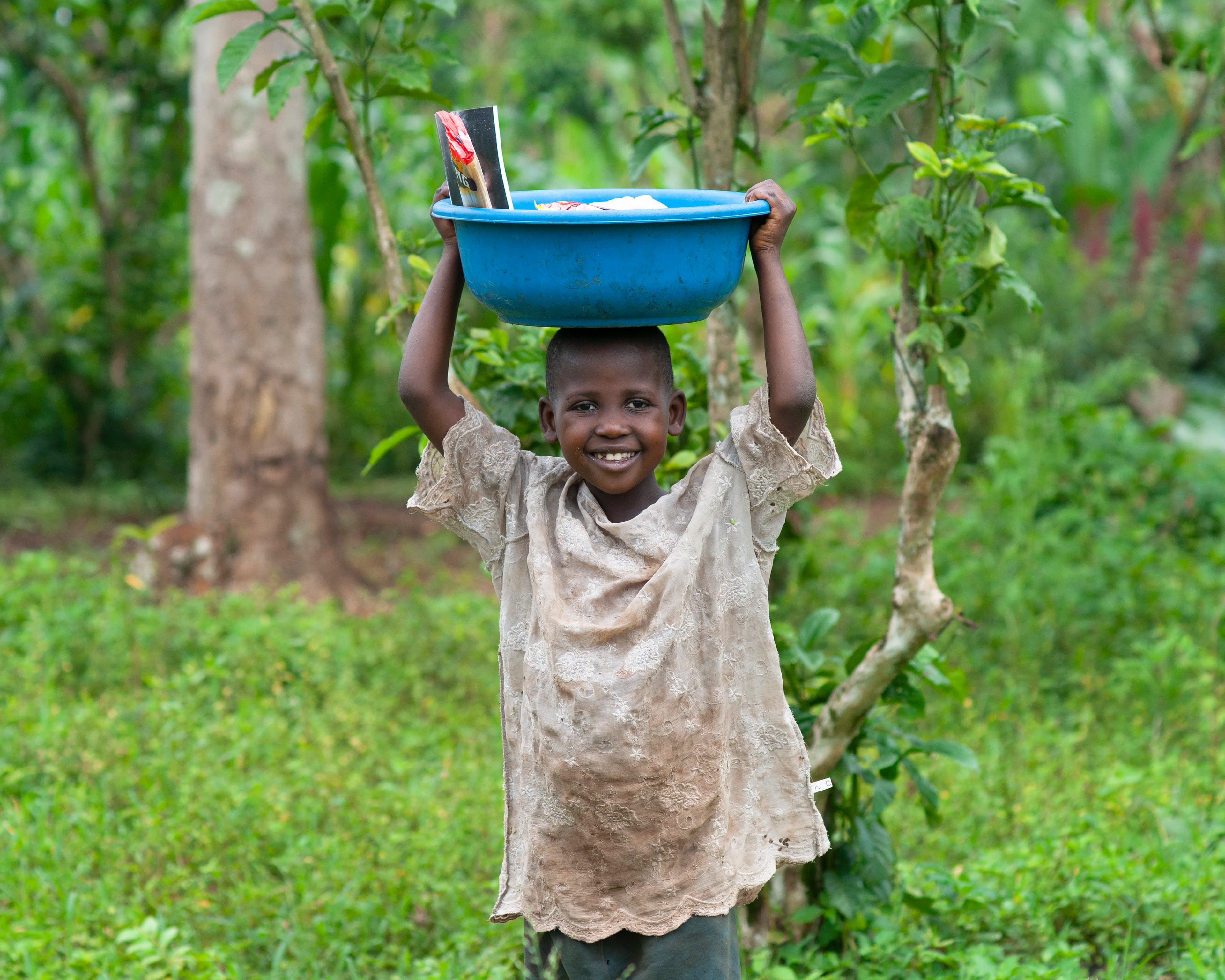 A child carries a blue basin of food on their head while standing among trees.