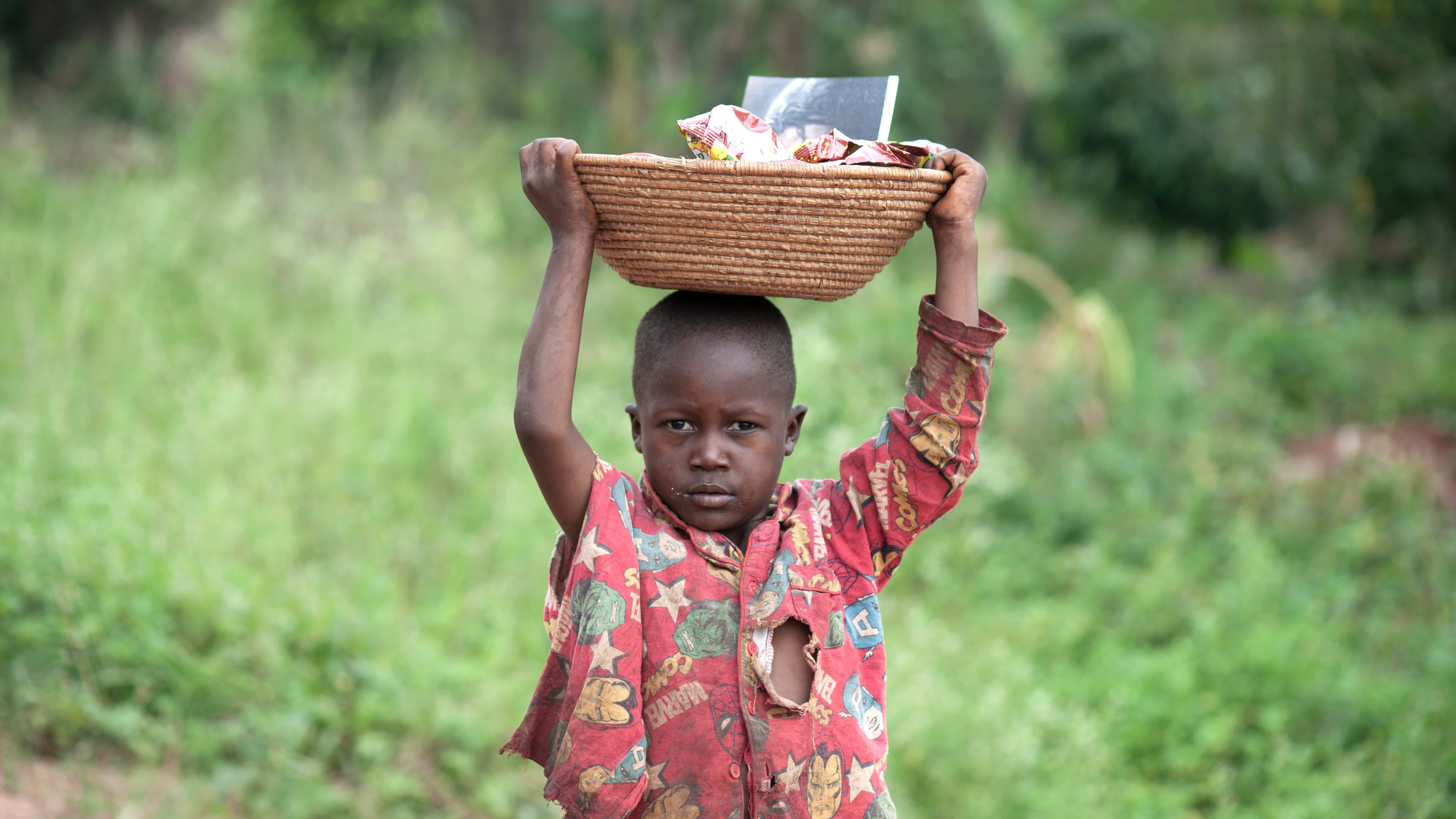 A child carries a basket of food items on their head while walking through greenery.