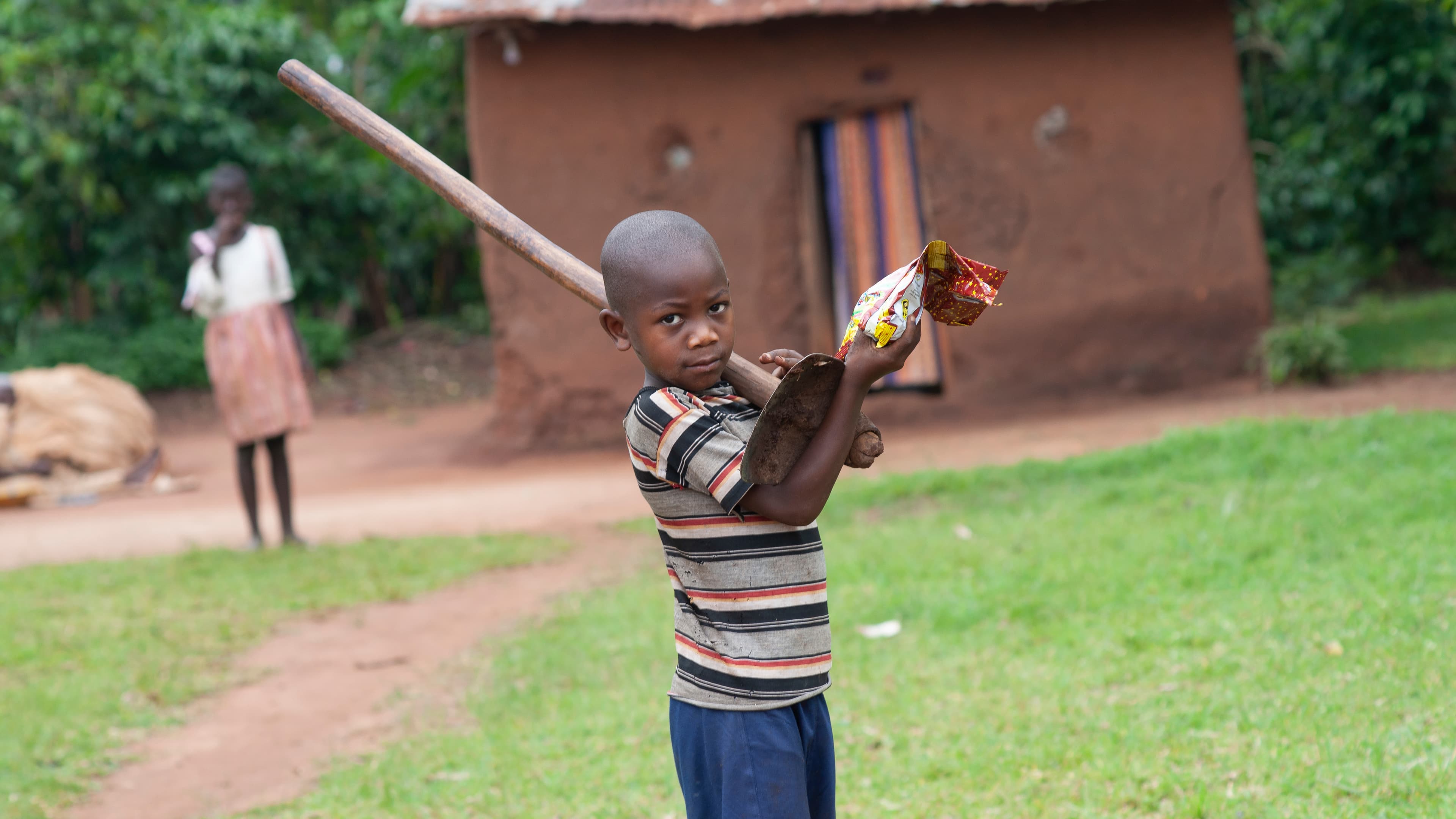 A boy holds a stick or tool while walking near a brick building, a woman visible in the background.