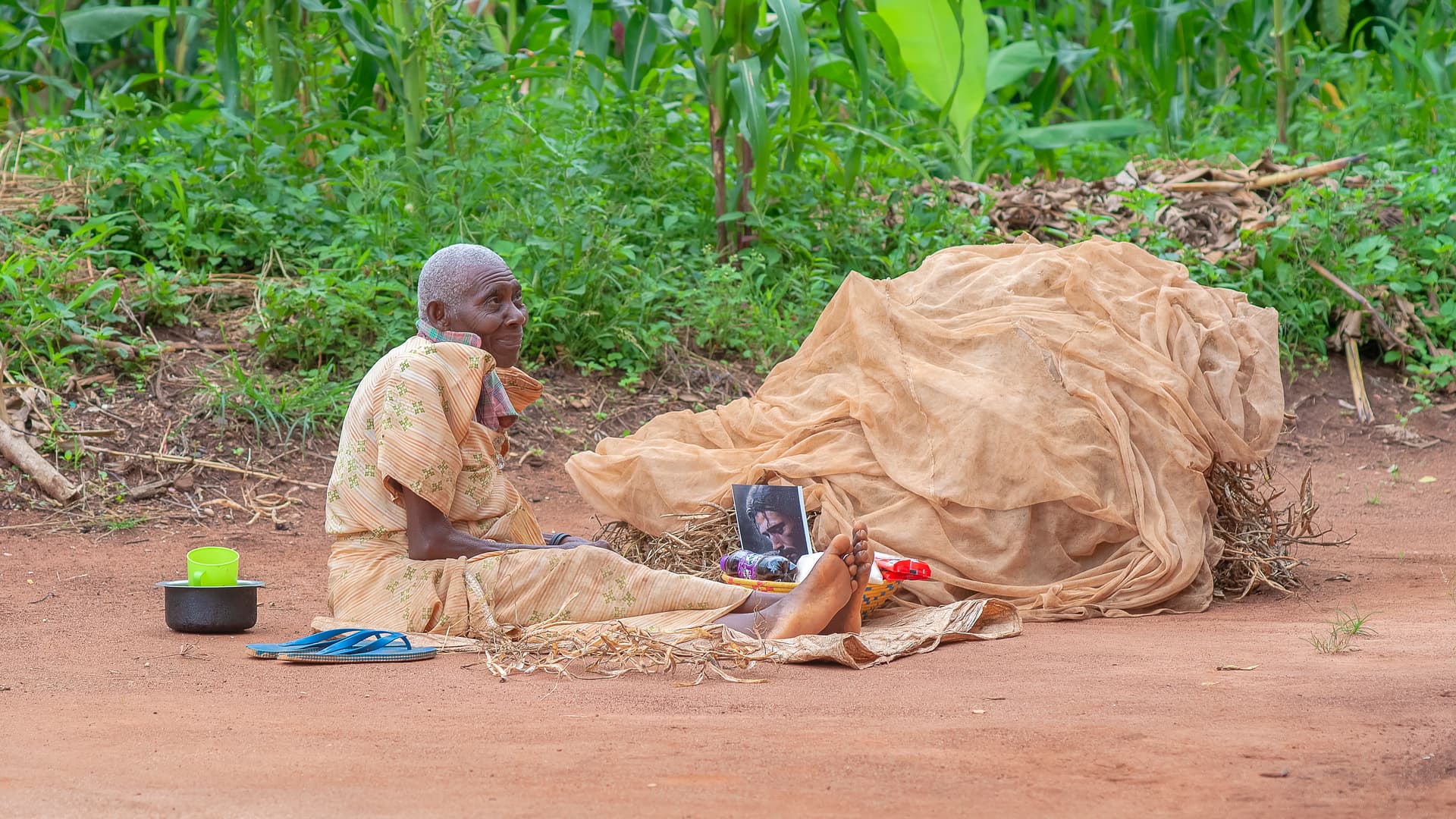 A person kneels beside someone lying on the ground covered by a woven mat, a food basin nearby.