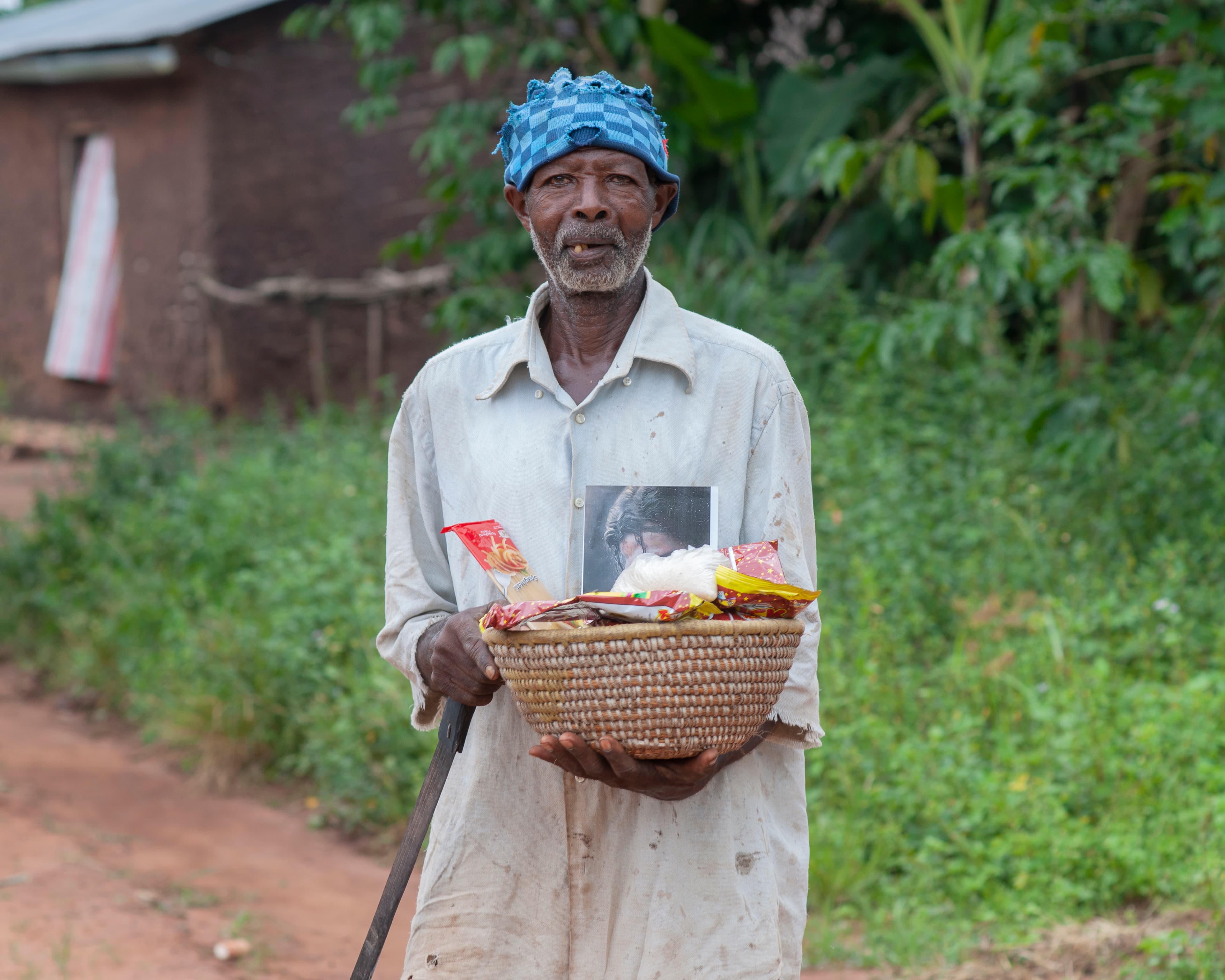 An elderly man in a blue headwrap holds a basket of food and a gospel tract outdoors.
