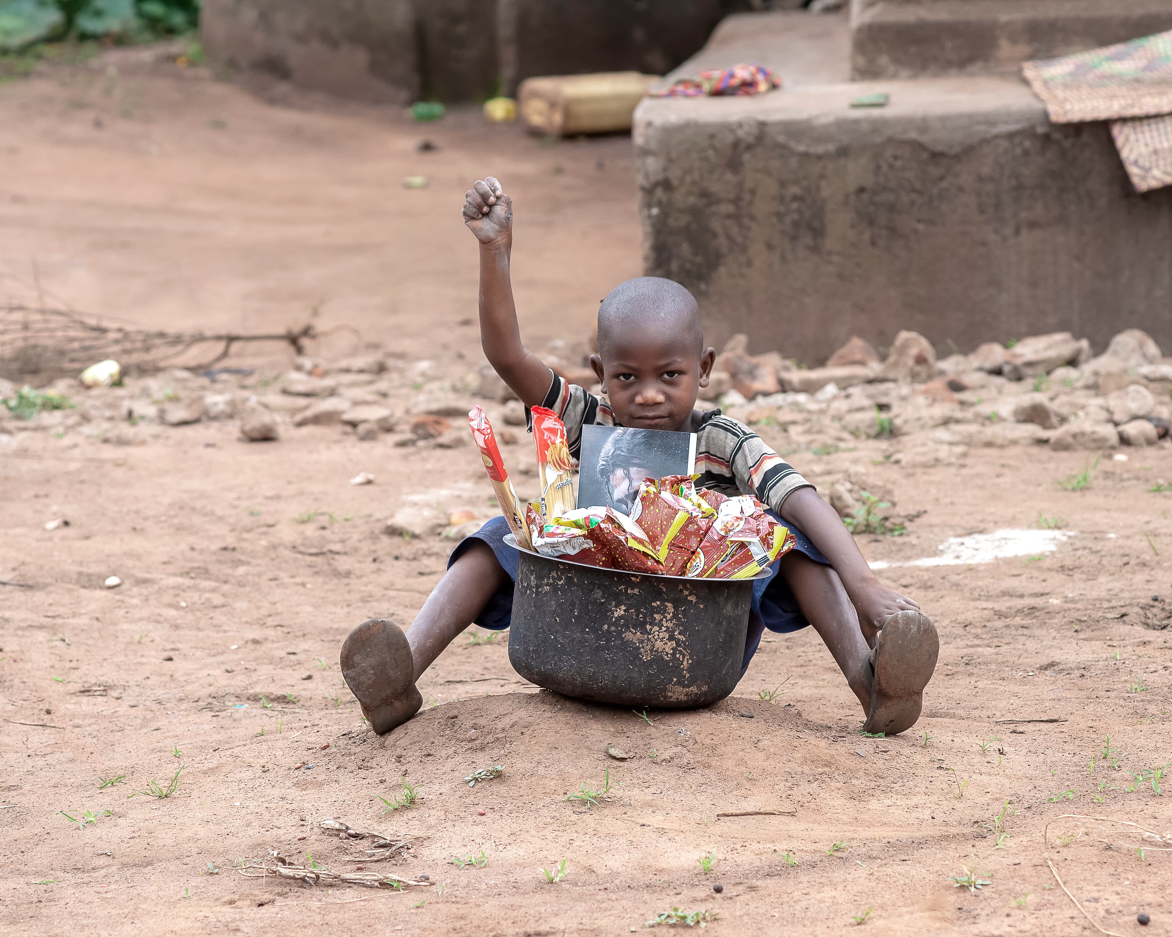 A toddler sits inside a metal basin outdoors on the dirt ground.