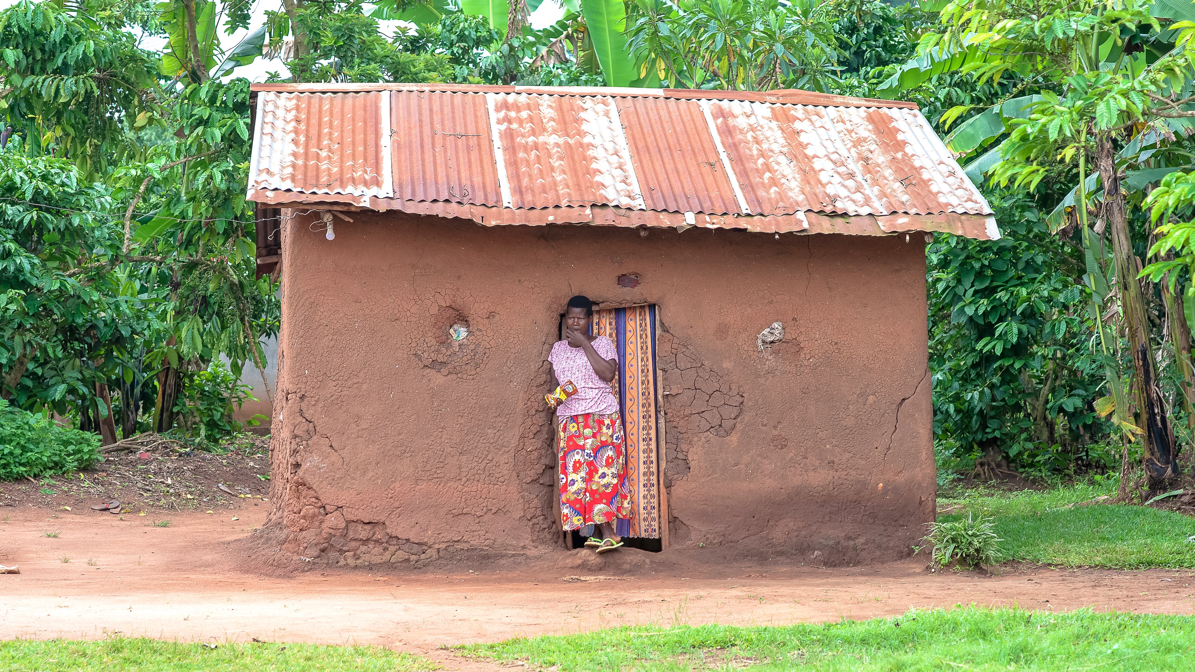 A small mud house with a corrugated metal roof, a woman standing in the doorway.