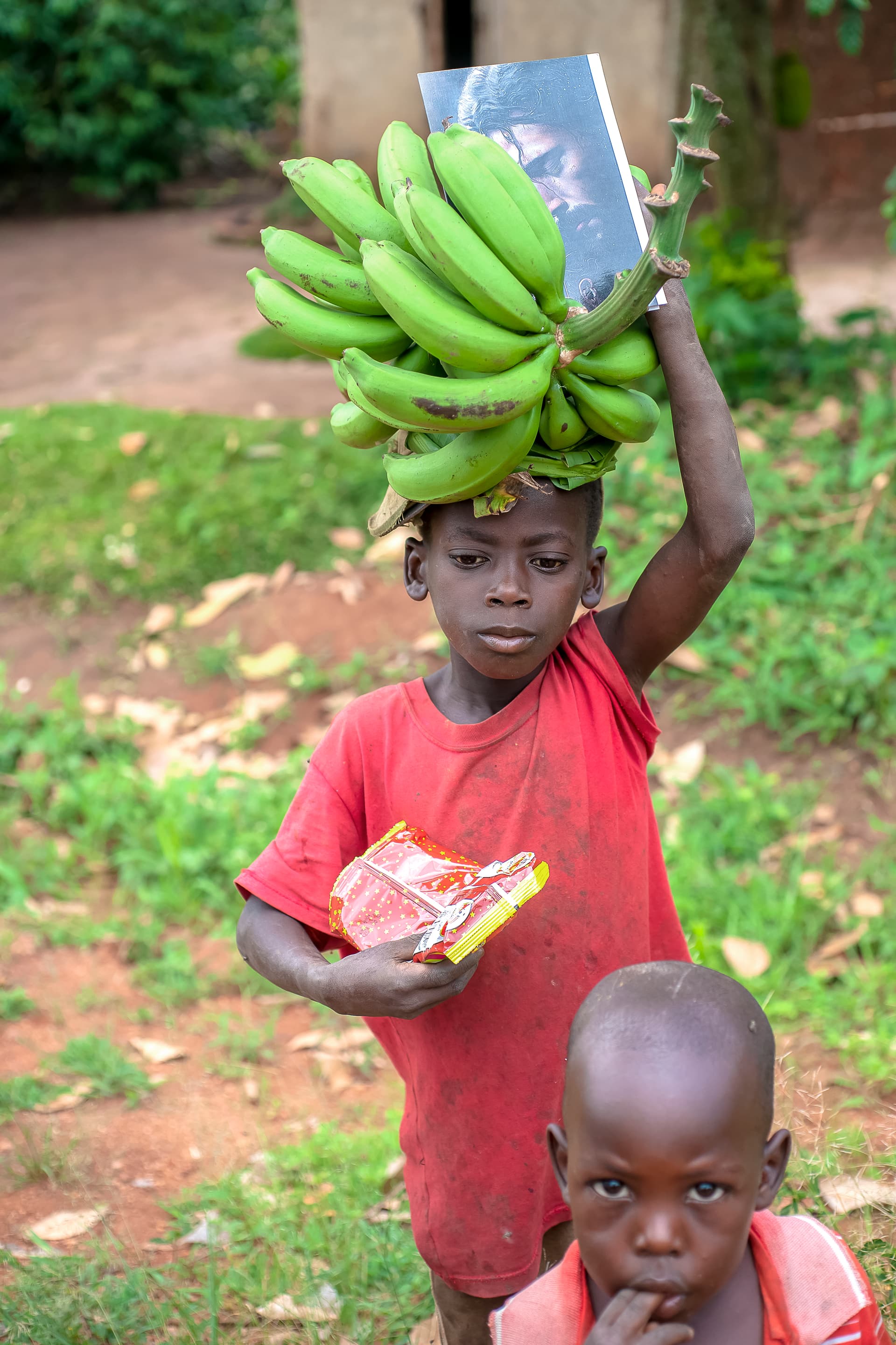 A girl carries a bunch of bananas and food items on her head with a younger child beside her.