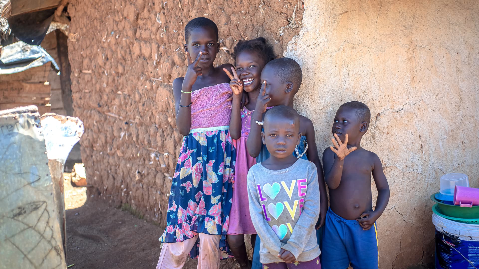 A group of children stand against a mud wall, a girl in a patterned dress making a peace sign.