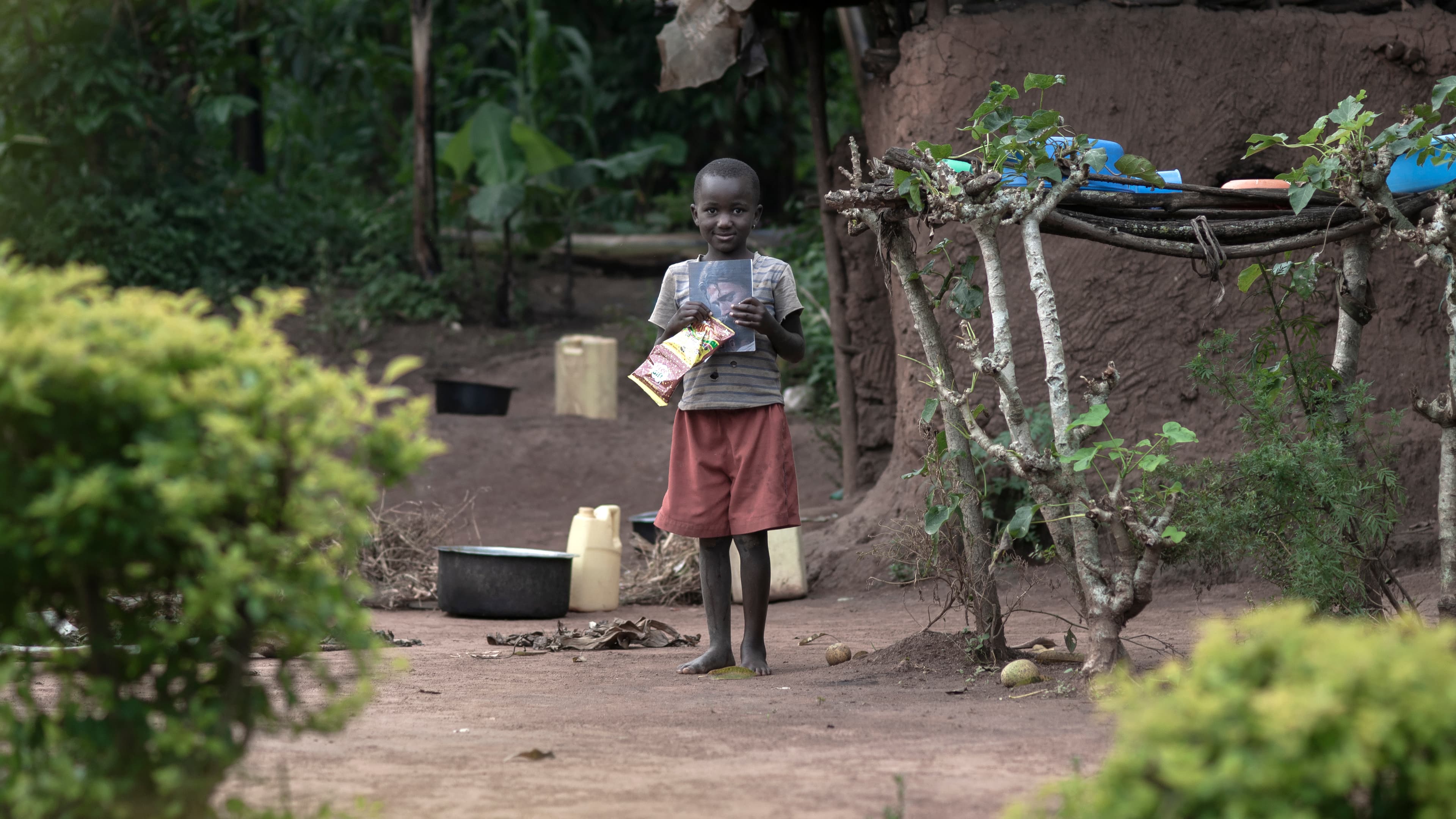 A child stands near shrubs and homes in the distance in a village setting.