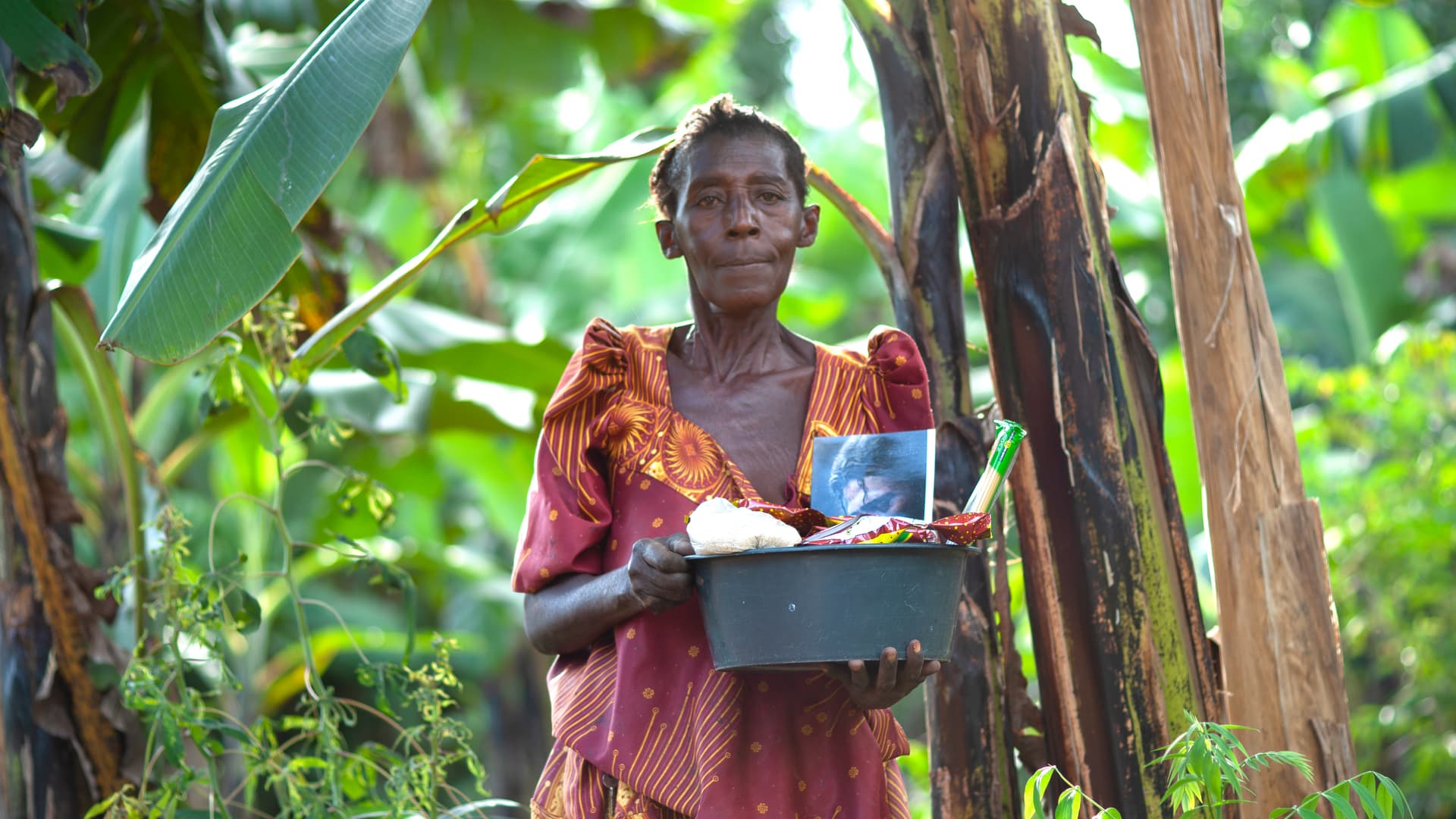 A woman carries a tray or basin of food items while standing among banana plants.