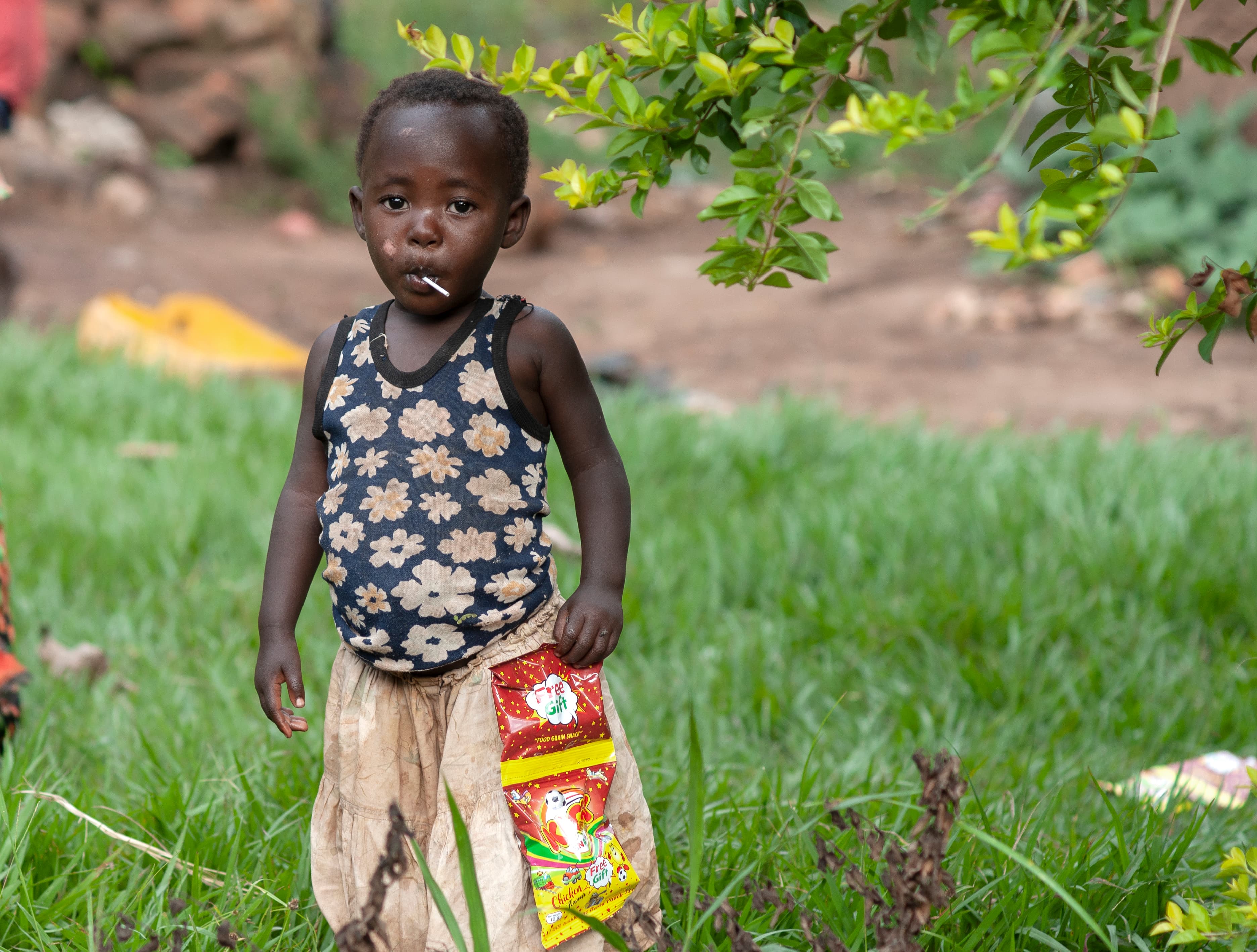 A child in a patterned outfit stands in grass holding a food gift bag.