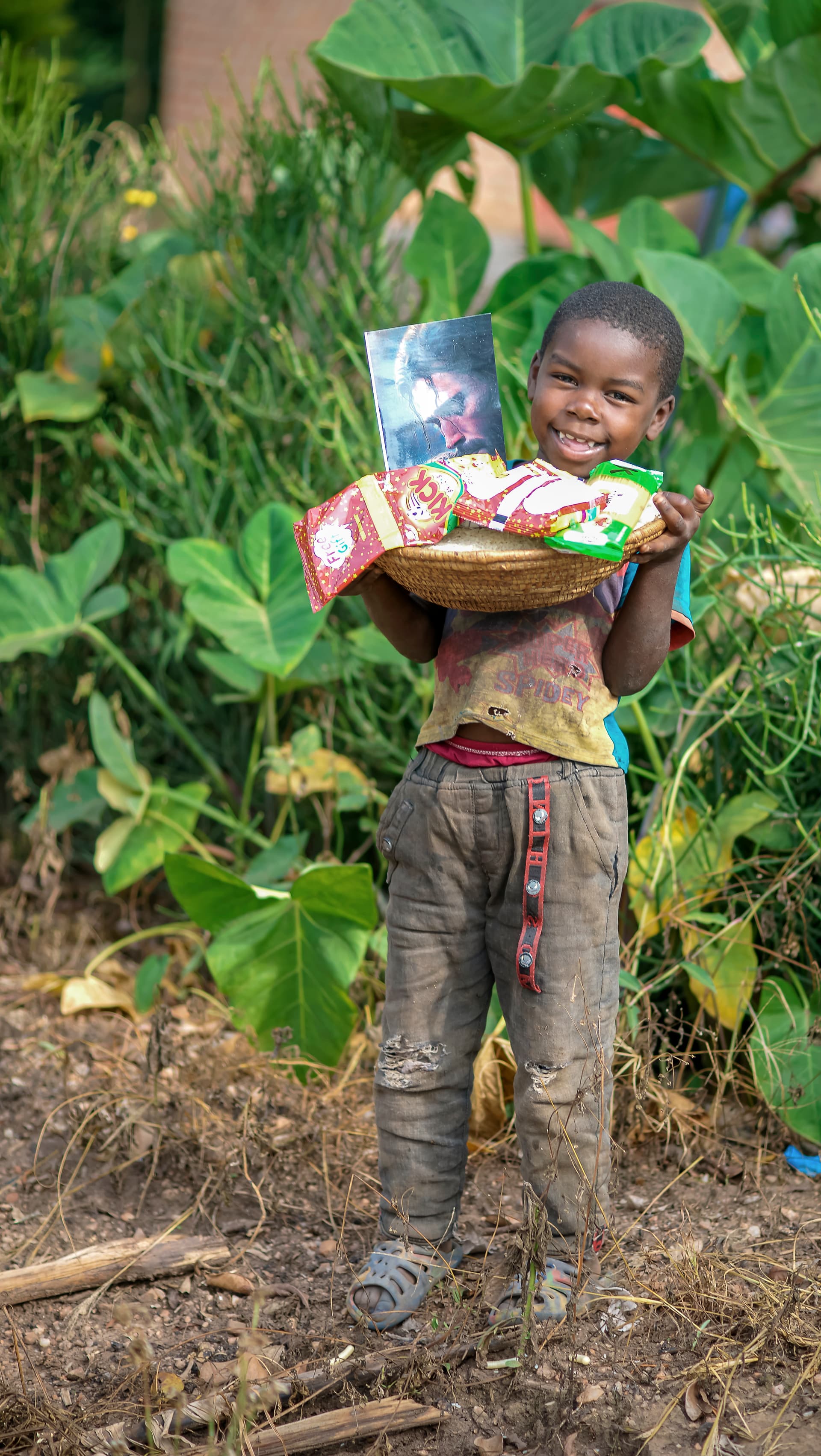 A boy carries a basket of food gifts on his head while standing among garden plants.