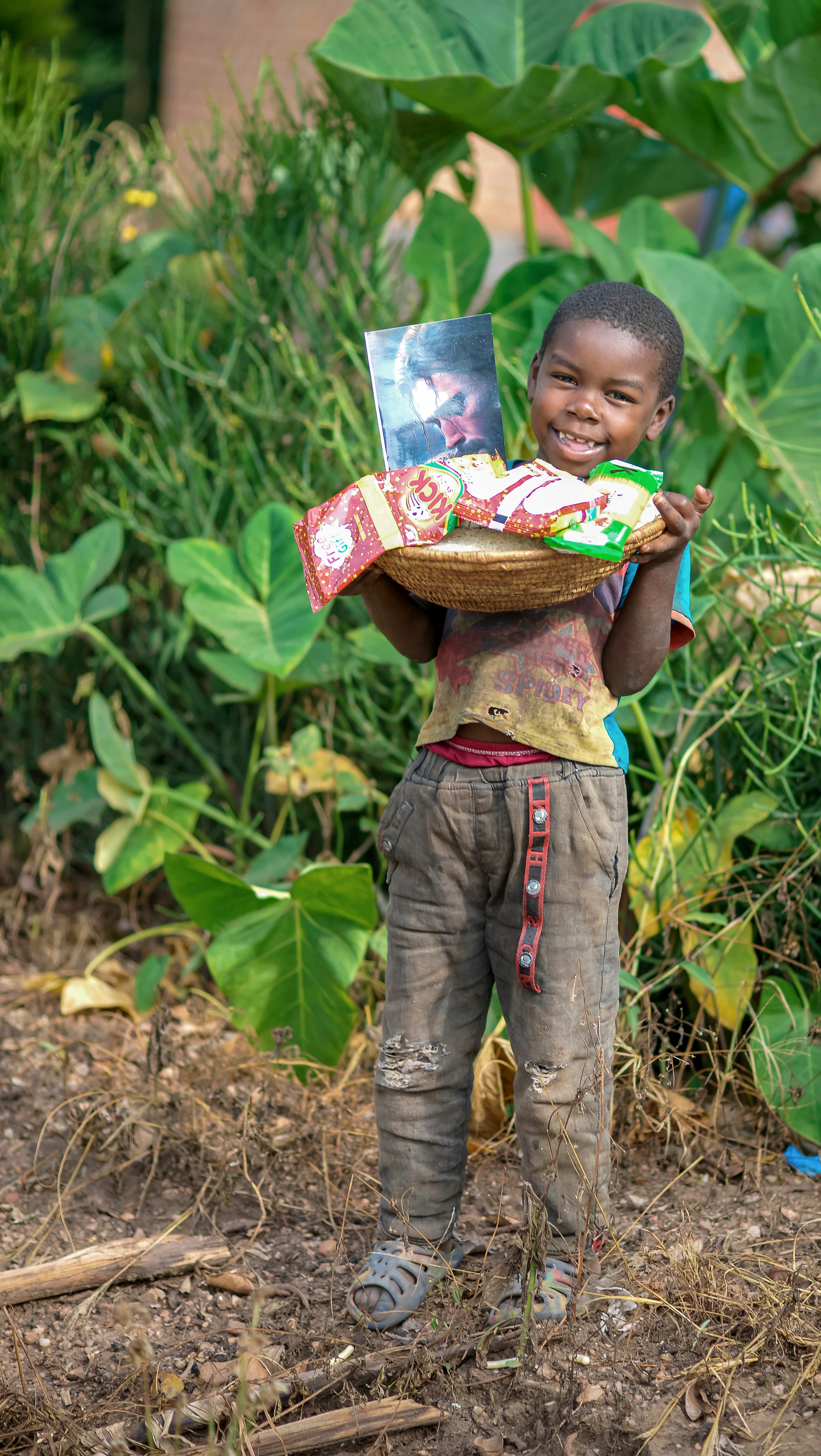 A boy carries a basket of food gifts on his head while standing among garden plants.