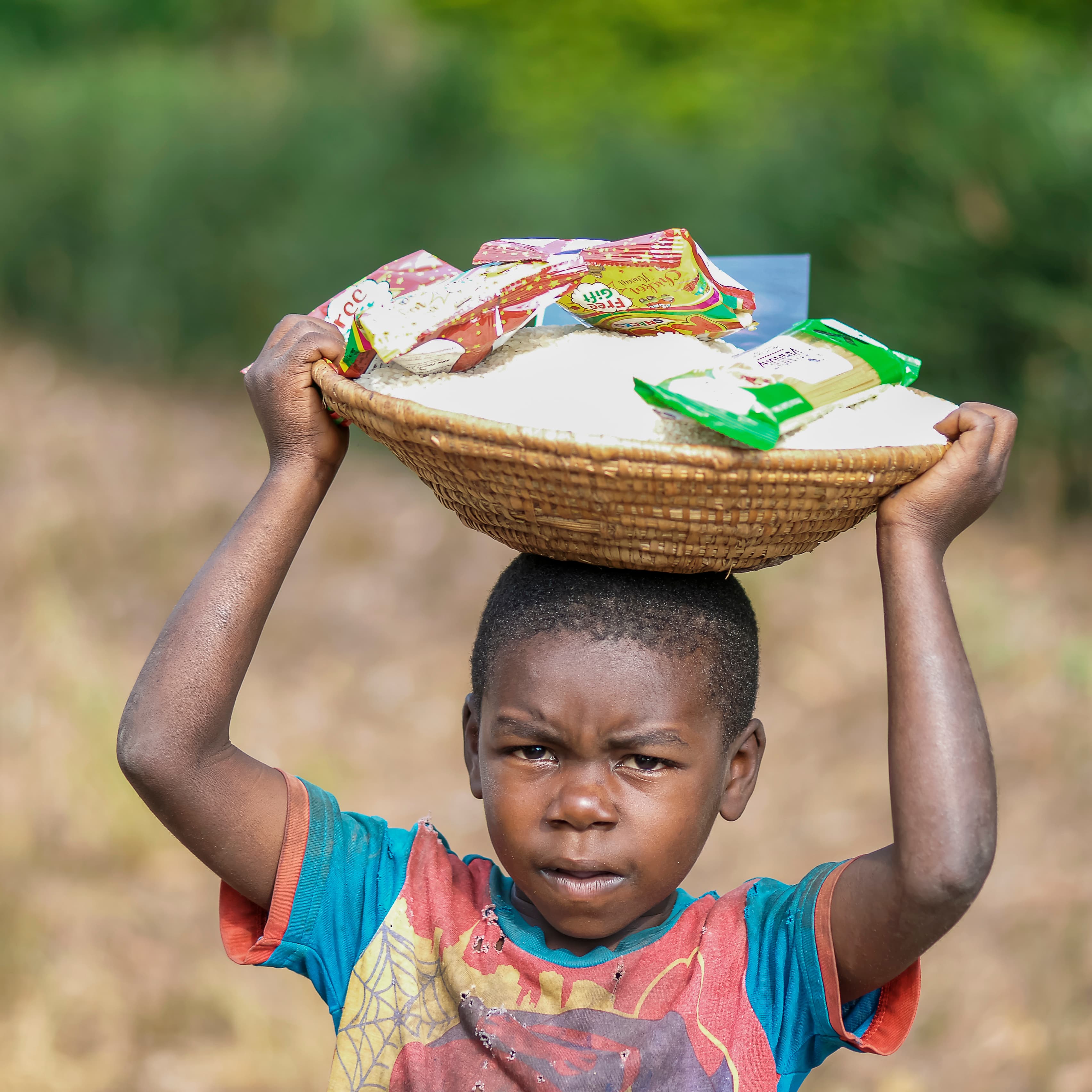A boy carries a woven basket of food and snack items balanced on his head, looking serious.