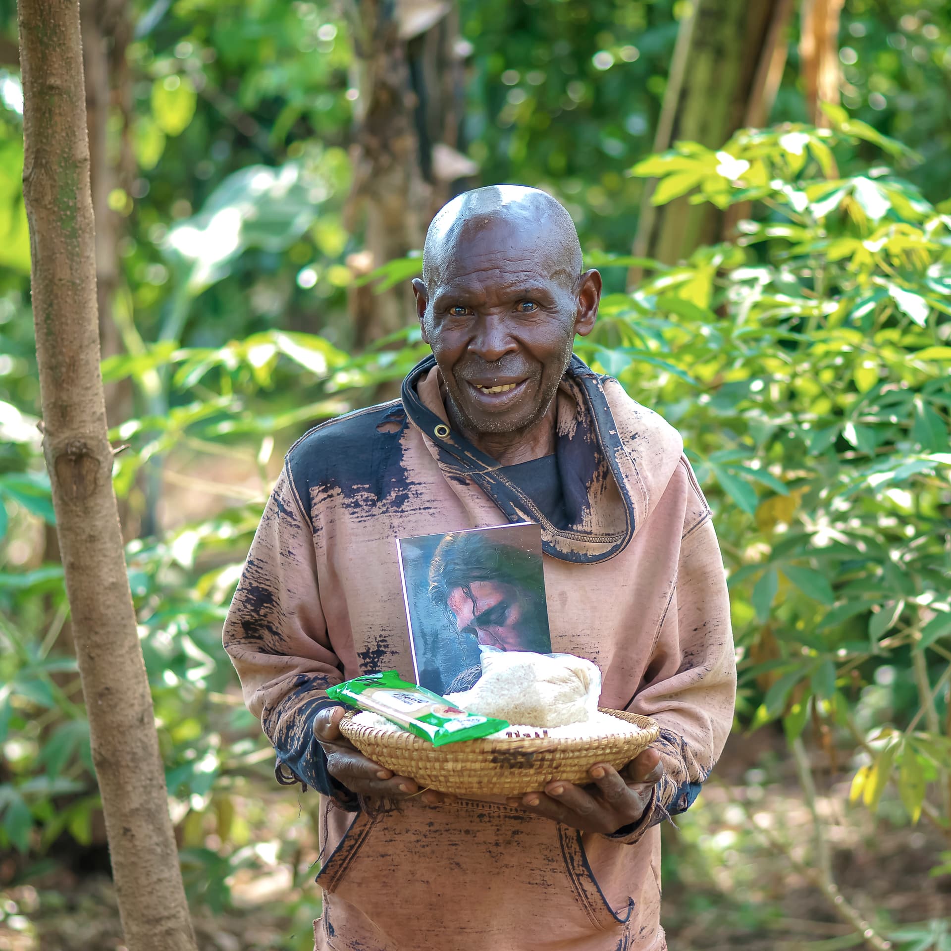 An elderly man holds a basket containing a photo and food items outdoors.