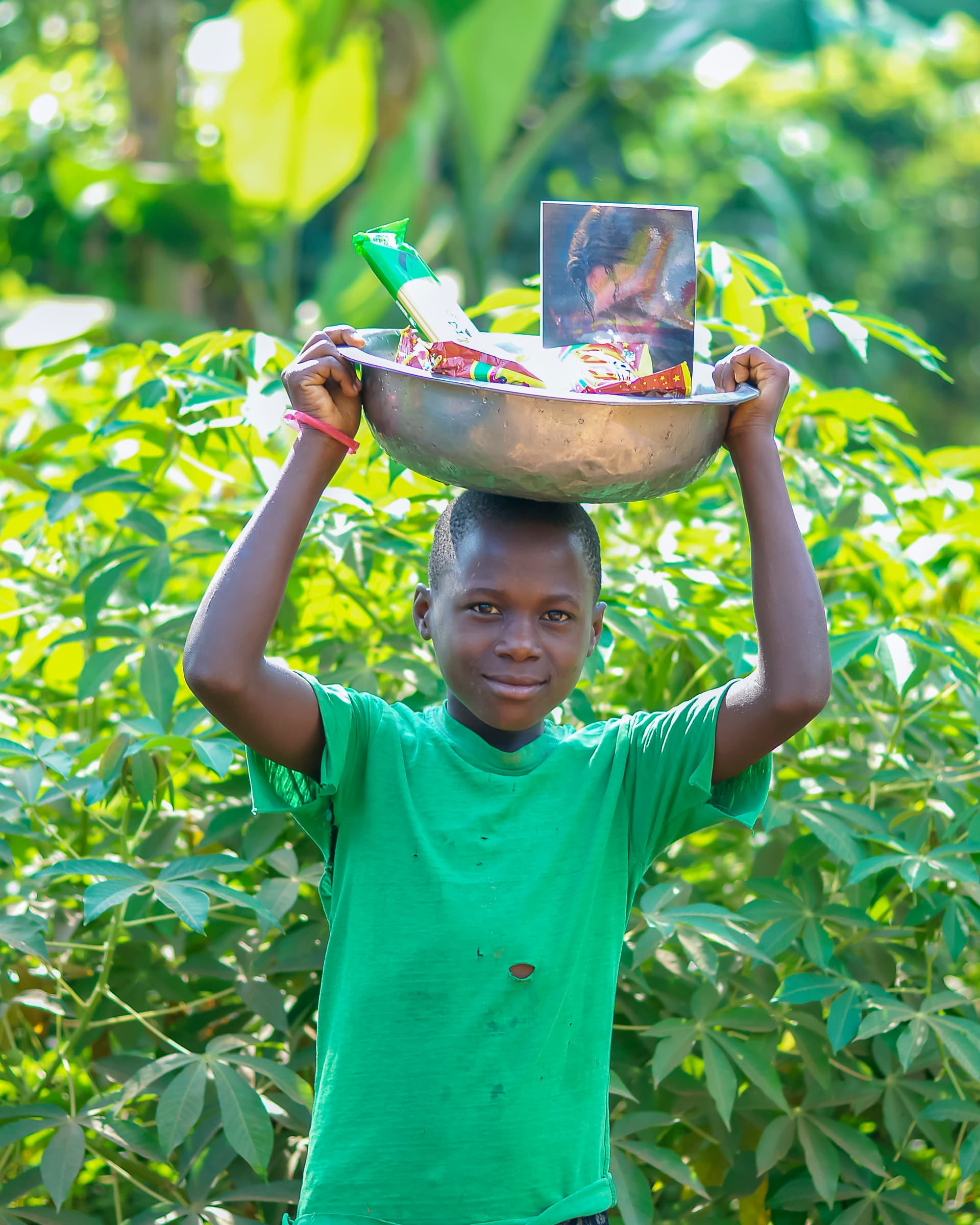 A boy in a green shirt carries a metal bowl with a photo and food on his head.
