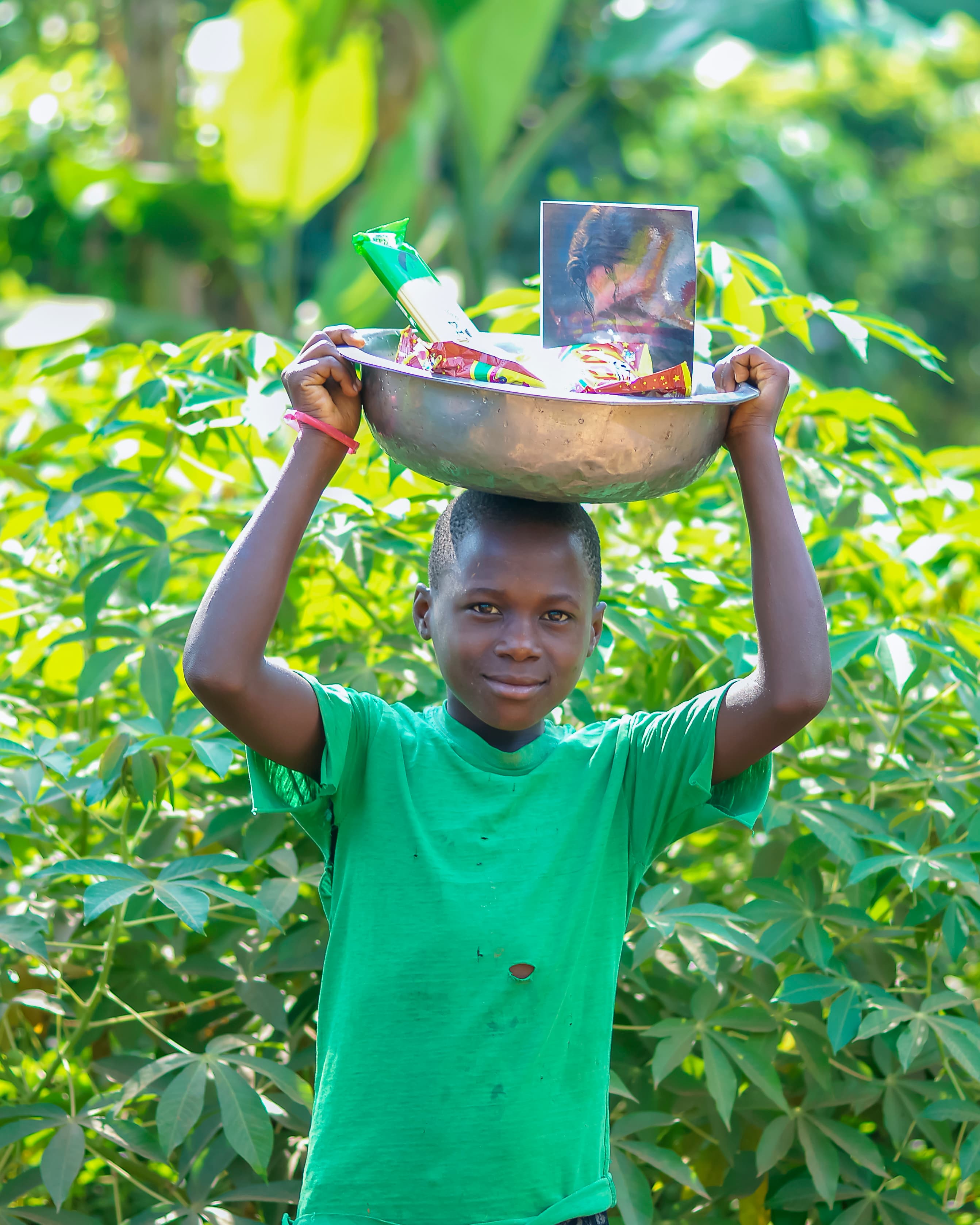 A boy in a green shirt carries a metal bowl with a photo and food on his head.