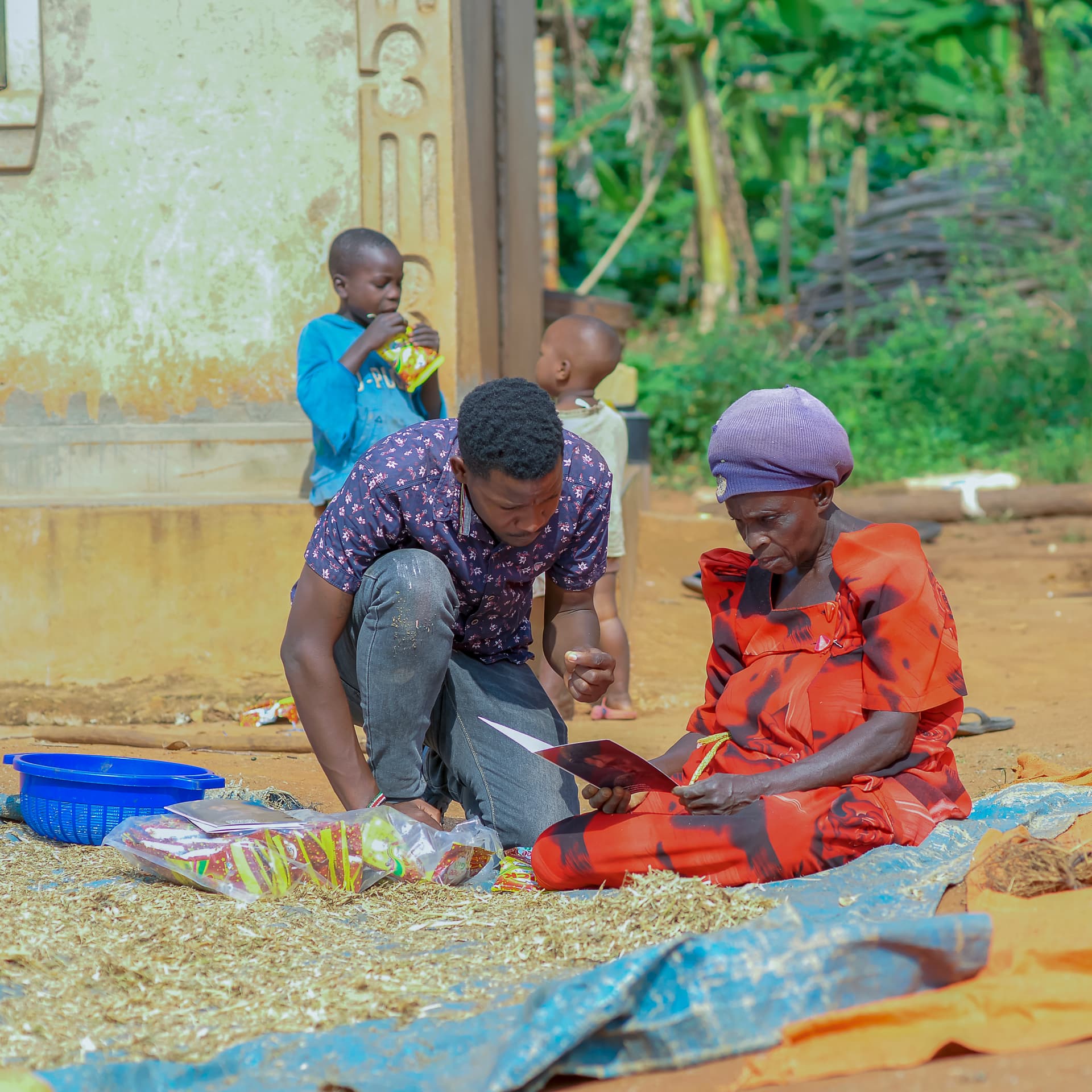 A man and woman kneel on the ground sorting dried maize kernels as a boy drinks from a bottle nearby.