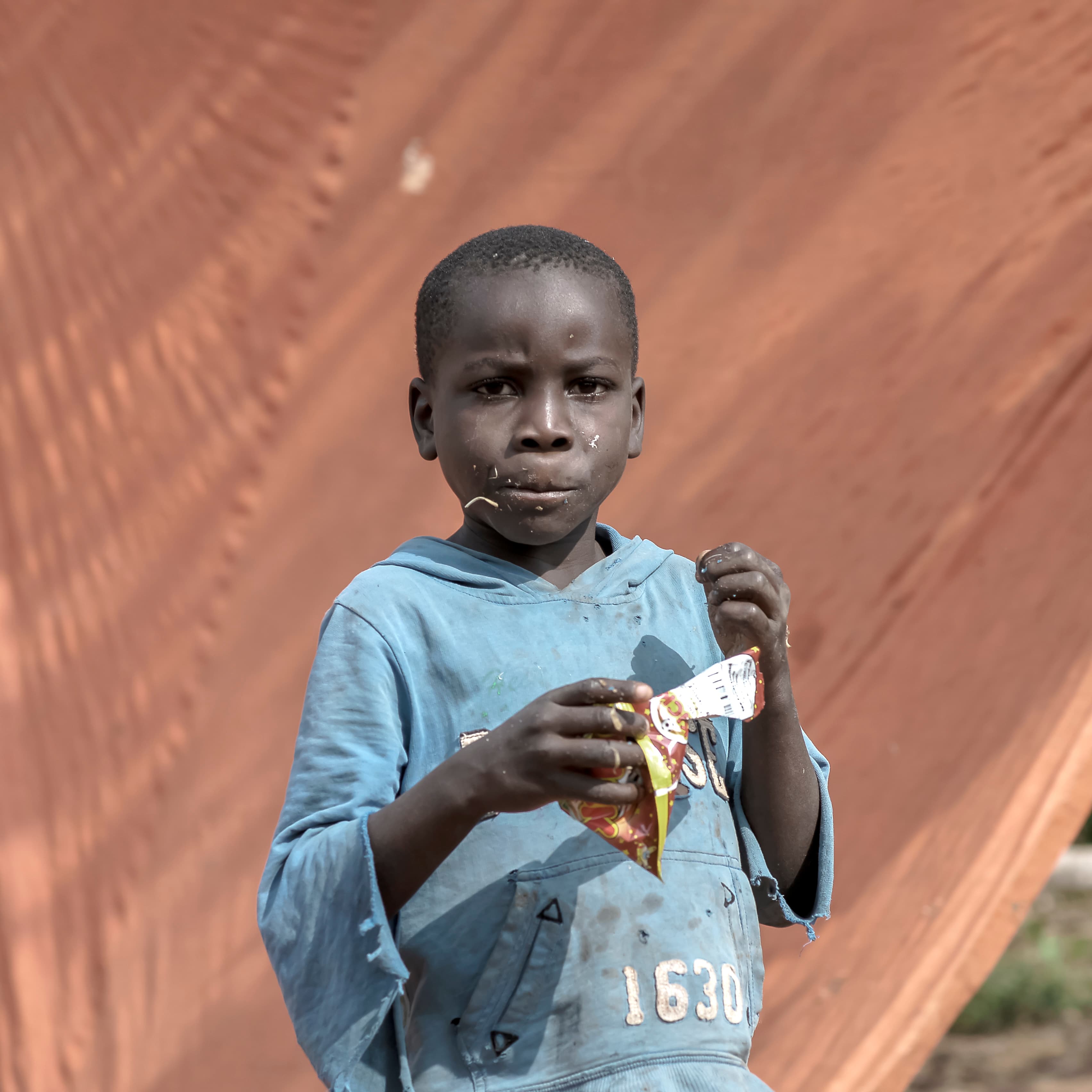 A boy in a blue-gray shirt holds a food packet against an orange-toned background.