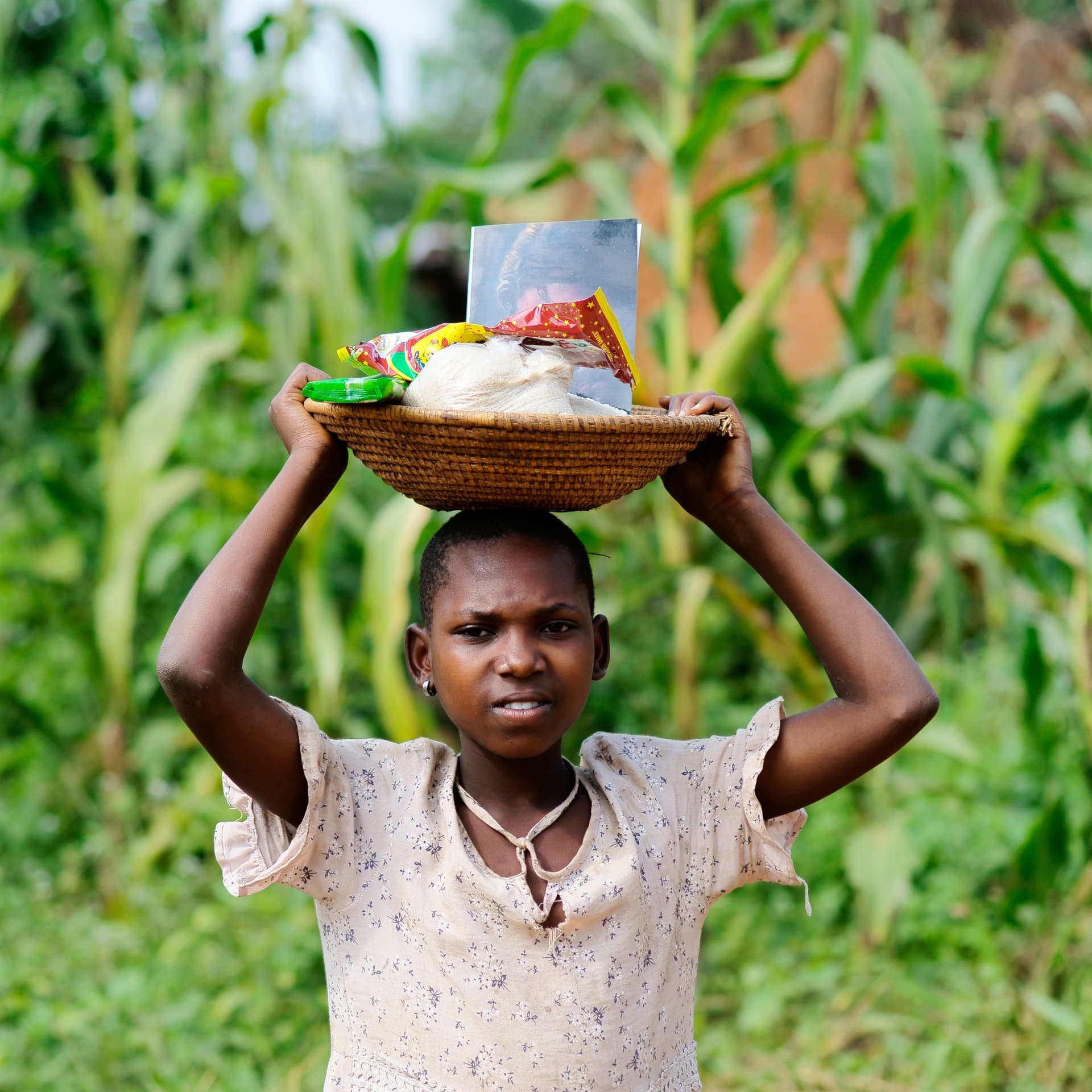 A woman carries a basket of food on her head while walking through tall grass.