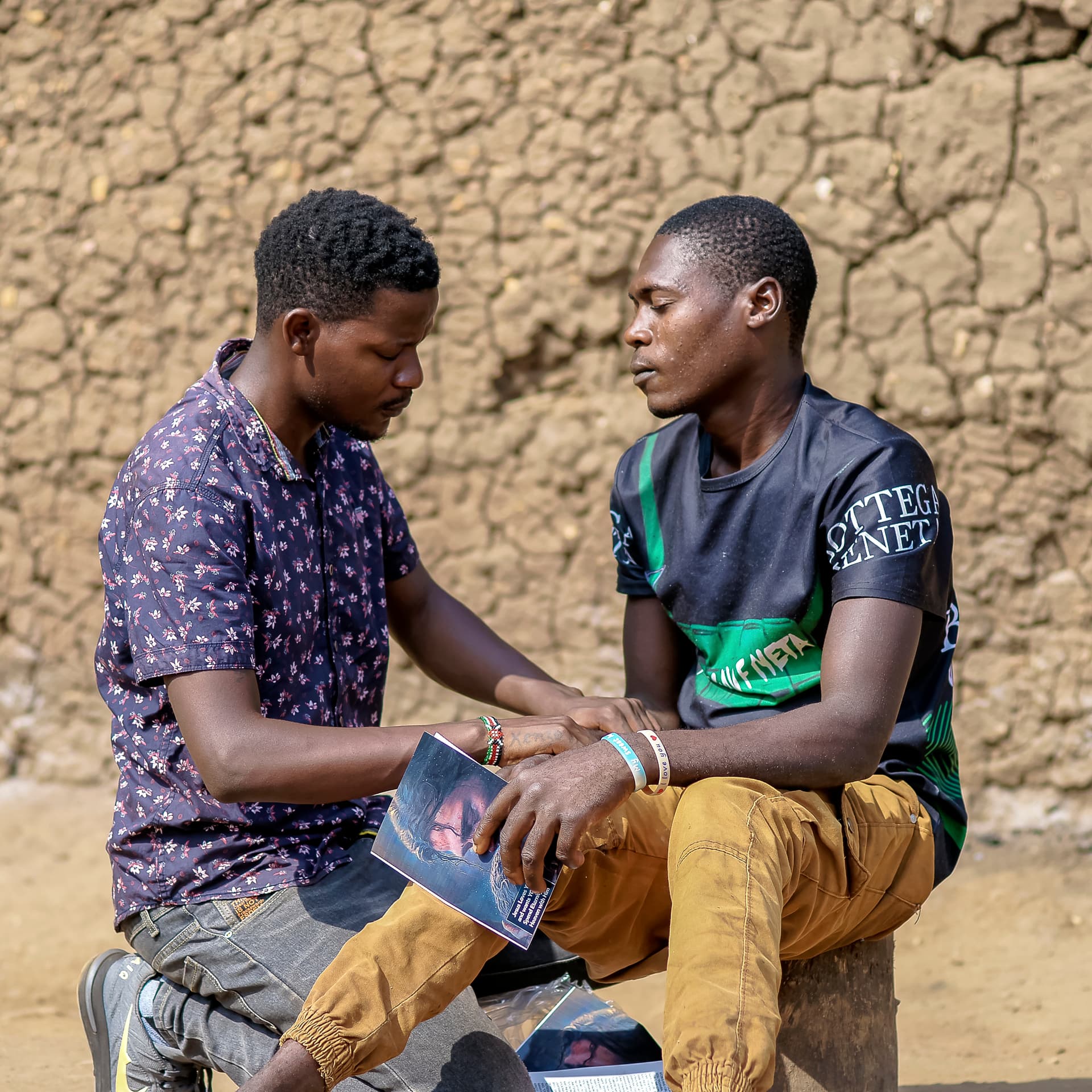 Two men sit together outdoors looking at a photograph or document.